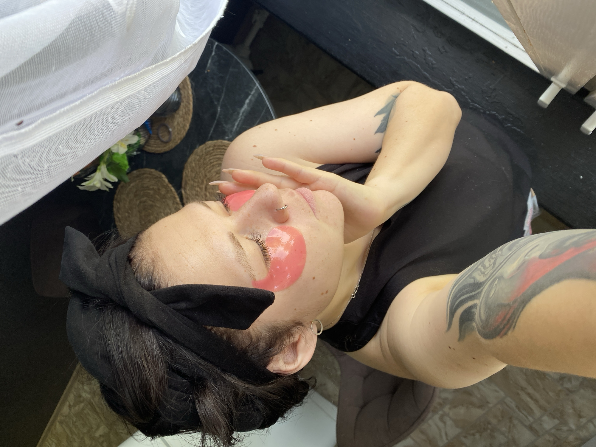 A high-angle selfie shows a person lying down. The individual has fair skin, dark hair restrained by a black headband, and is wearing pink facial patches under each eye. The person's hands are near their face, with long fingernails visible. Tattoos are present on both arms. The person wears a black top.
The background includes a black marble-look table with straw placemats, white curtains, and flowers in a vase. Through a window, an outdoor view suggests a possible balcony setting. This scene purportedly occurs in Chaikovsky, Russia, though the image alone doesn't provide definitive confirmation of that location.