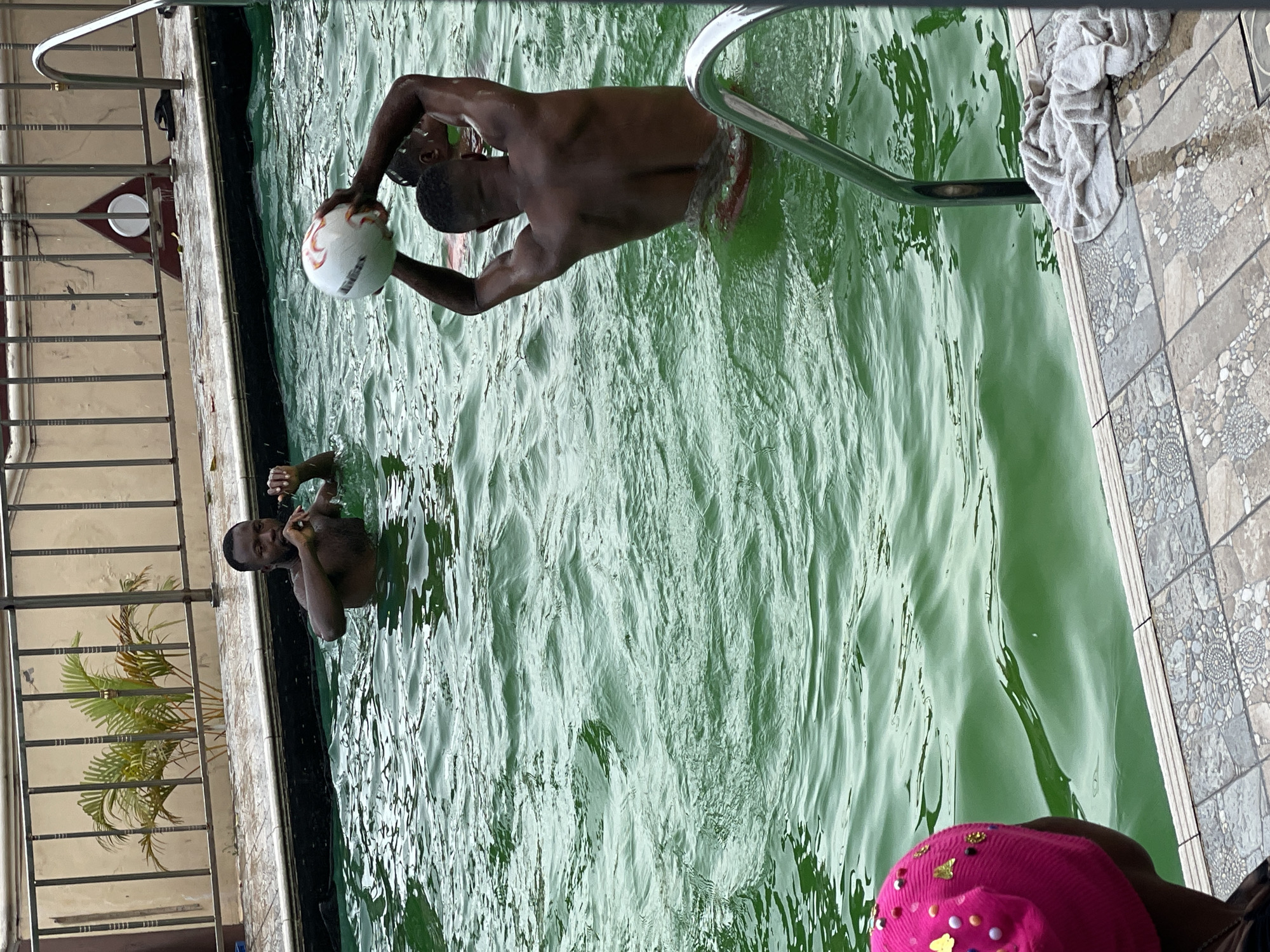 In an outdoor swimming pool filled with green water, three individuals are visible. One person is standing in the water up to their chest, holding a white and red striped ball above their head. Their back is to the camera. To the left of this person, another individual is partially submerged in the water, looking towards the person with the ball. A third individual, wearing a bright pink beanie adorned with colorful embellishments, is partially visible in the foreground on the left, facing away from the camera and looking into the pool. The pool area is bordered by patterned tiles and a metal railing. A towel is draped over a metal pool ladder. In the background, a building with a beige wall and a metal fence is visible, with a palm plant behind the fence.