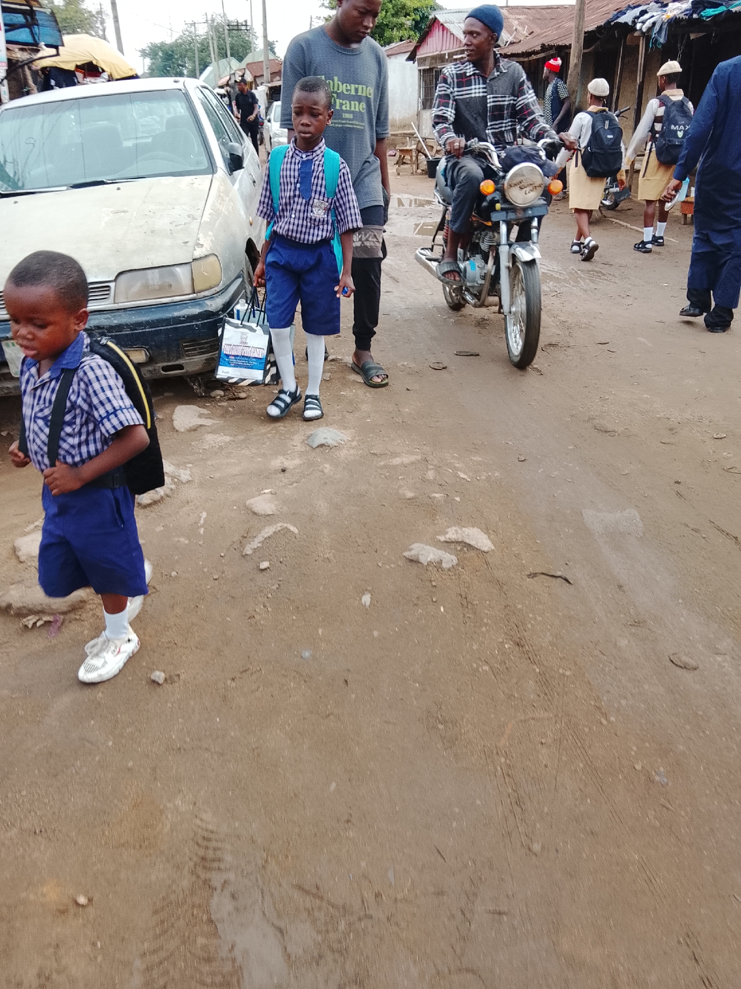In Abuja, Nigeria, a dirt road depicts everyday life. A young child in a blue school uniform walks with a backpack, seemingly towards the left of the frame, while another stands near a worn, white car. A man wearing a grey shirt with the text "Taberne Frane 1965" stands behind the second child. To the right, a man rides a motorcycle, with other pedestrians, some in school uniforms, nearby. Buildings with corrugated metal roofs line the background.