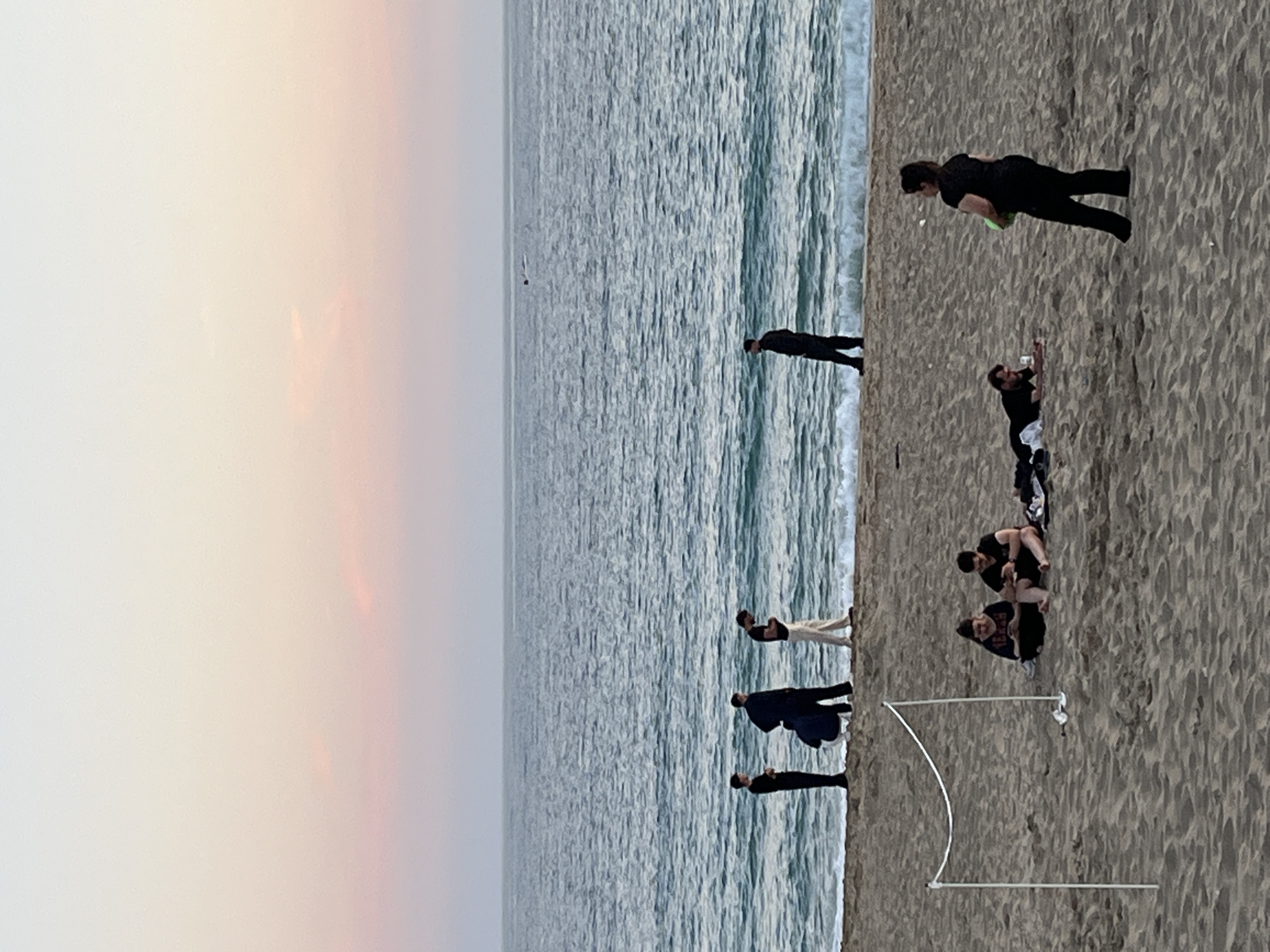 The image depicts a beach scene with several people present. The sky transitions from a light peach color near the horizon to a muted gray. The ocean occupies a significant portion of the image, showing gentle waves. On the sandy beach, there are multiple individuals engaged in various activities: one person is bent over as if picking something up, others are standing or sitting. One structure is a white curved object which could be an event border. The location is inferred to be Alanya, Turkey, since it was specified.