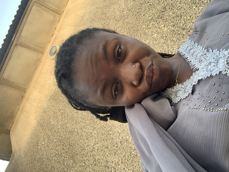 A headshot selfie, captured in Bojude, Nigeria, depicts an individual against a textured light-brown wall. The person, appearing to be an adult female with dark skin and dark hair styled in braids or dreadlocks, faces forward, making direct eye contact with the camera. She has a small, shiny stud piercing in her left nostril, and her lips are slightly pursed. She wears a grey garment featuring a ribbed texture, white lace trim around a V-neckline, and small, shiny embellishments. A thin gold-colored chain necklace is visible, and a light grey fabric is draped over her left shoulder. In the upper background, a portion of a building's eaves or roof overhang, constructed from light-colored panels, is visible, along with a circular wall-mounted light fixture on the wall. The scene is brightly illuminated by natural light.