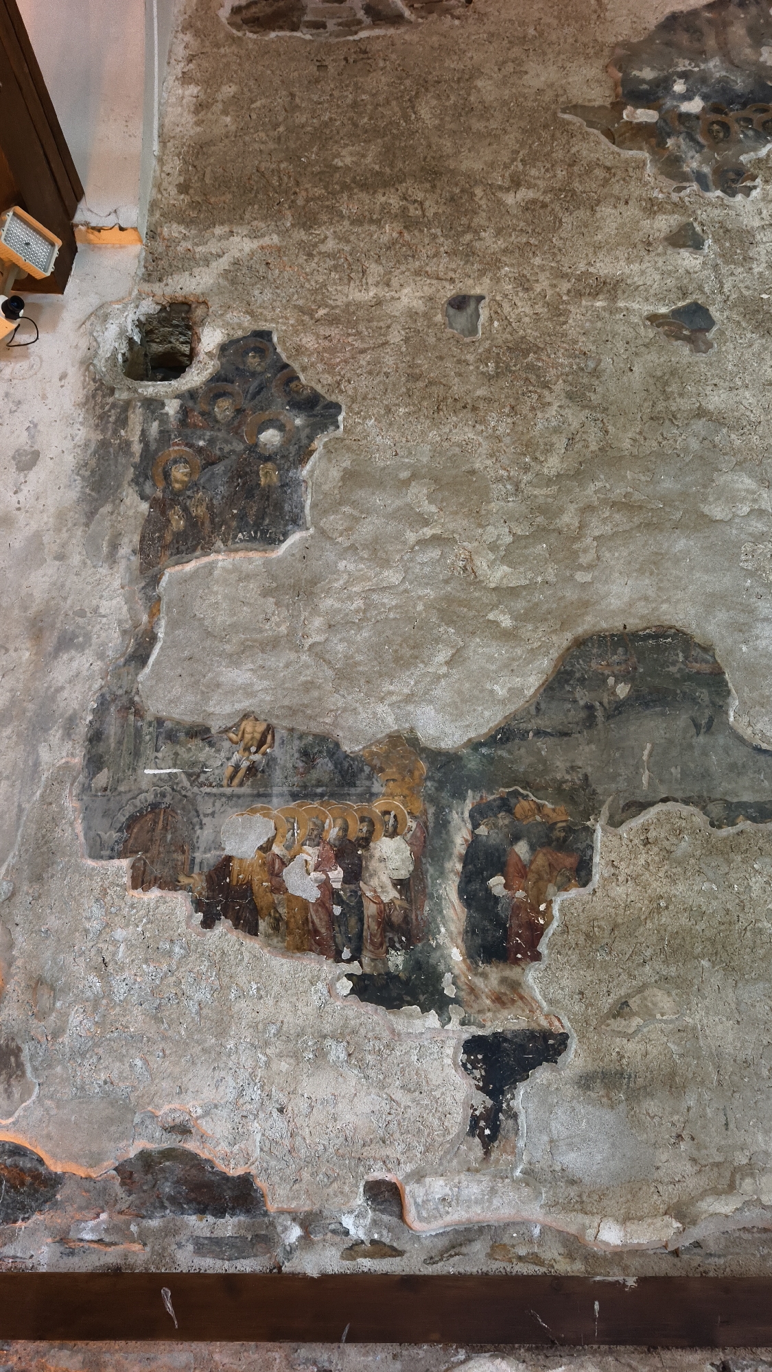 This image depicts a section of a fresco on a weathered stone wall in the city of Selçuk, Turkey. The fresco is significantly damaged and fragmented, with large areas of plaster missing, revealing the rough stone beneath. What remains of the painting depicts several figures, some with halos, suggesting a religious or historical scene. The figures appear to be stylized and rendered in muted colors, with earthy tones dominating. In the upper left portion of the fresco, there are circular elements, possibly halos, and fragmented human forms. Below this, a larger section of the fresco reveals a group of figures standing together, with some individuals depicted with more detail, including clothing and facial features. There's a figure with a lighter complexion in the center of this group. To the right of this group, and slightly higher, another fragmented depiction shows figures. The overall impression is one of historical decay and the remnants of ancient artistry. The wall itself is rough and uneven, with areas of plaster showing different textures and colors. Light fixtures, likely for illumination, are visible in the upper left corner of the frame, casting a warm glow on the wall.