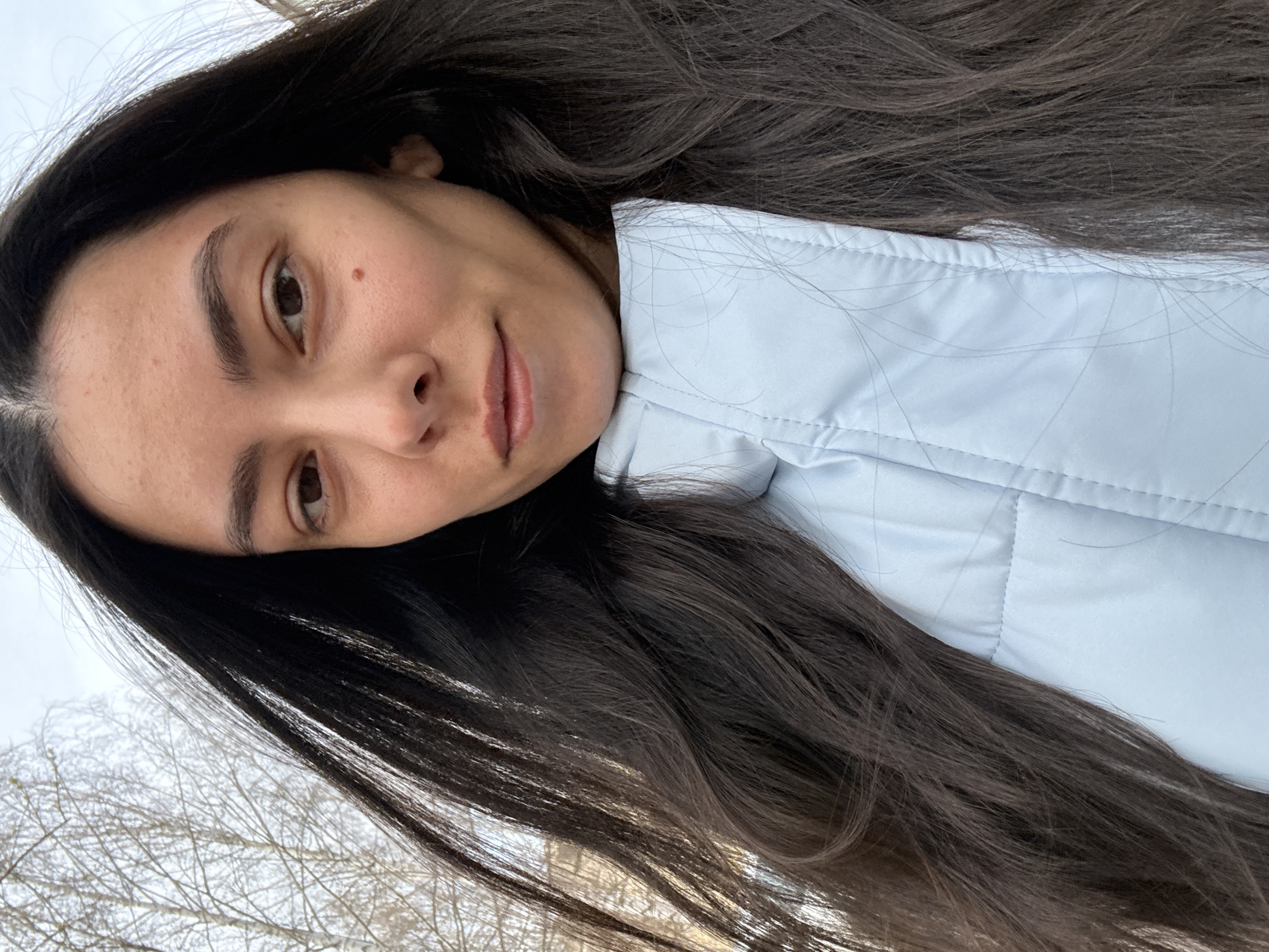 A close-up, portrait-style image captures a young woman outdoors. Her head is tilted to the side as she looks directly at the camera with a gentle, calm expression. She has long, dark brown hair that cascades over her shoulders and partially covers a light blue, possibly padded jacket or coat, characterized by visible horizontal stitching and a structured collar. Her facial features include dark eyes, well-defined eyebrows, and a distinct small mole situated above her left eyebrow. Her lips are naturally colored and curved in a subtle, soft smile.
The background is softly blurred, revealing a bright, diffused sky that suggests overcast conditions, and a dense network of bare tree branches, indicative of late winter or early spring. This outdoor setting is consistent with the provided location context of Chaikovsky, Russia. The lighting is even and natural, suggesting it is daytime. There is no visible text in the media file.