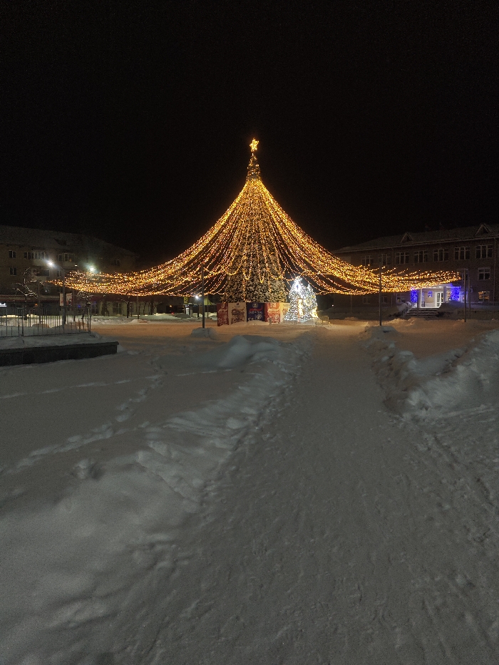 A wide snowy path leads toward a central Christmas tree. Draped in warm, golden lights, the tree is adorned with a star on top. A smaller, illuminated white tree stands nearby. Colorful wrapped "gifts" surround the base of the larger tree. Buildings with lit windows flank the scene in the background. The dark sky suggests it is nighttime.