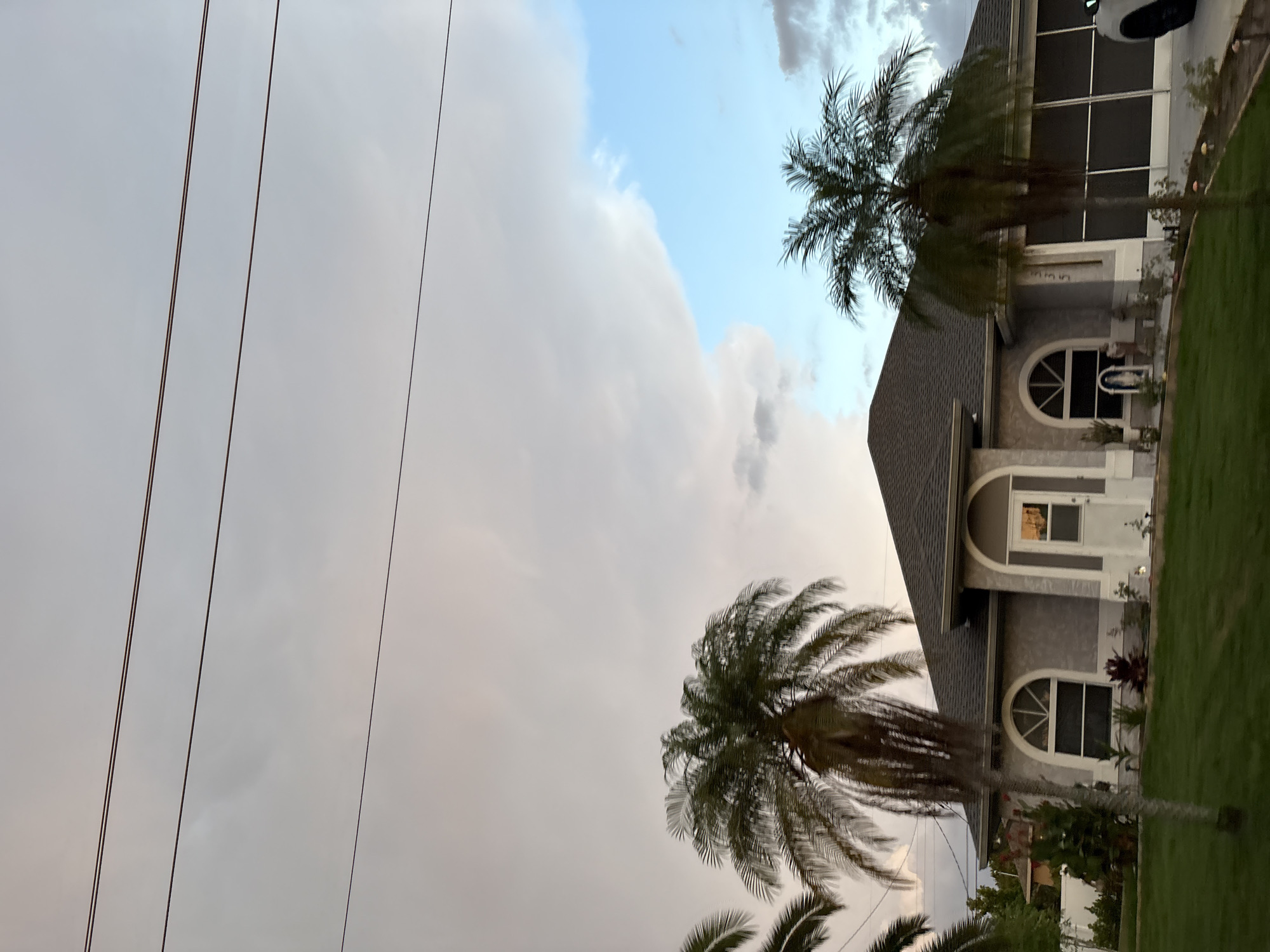 In the city of South Venice, United States of America, a single-story house with a grey exterior and a dark grey roof is visible. The house features arched windows and a white front door. Two palm trees stand in front of the house, with one on the left and another on the right. The sky above is filled with clouds, with a patch of blue sky visible on the right side. Power lines are strung across the top of the image. The lawn in front of the house is green and well-maintained.