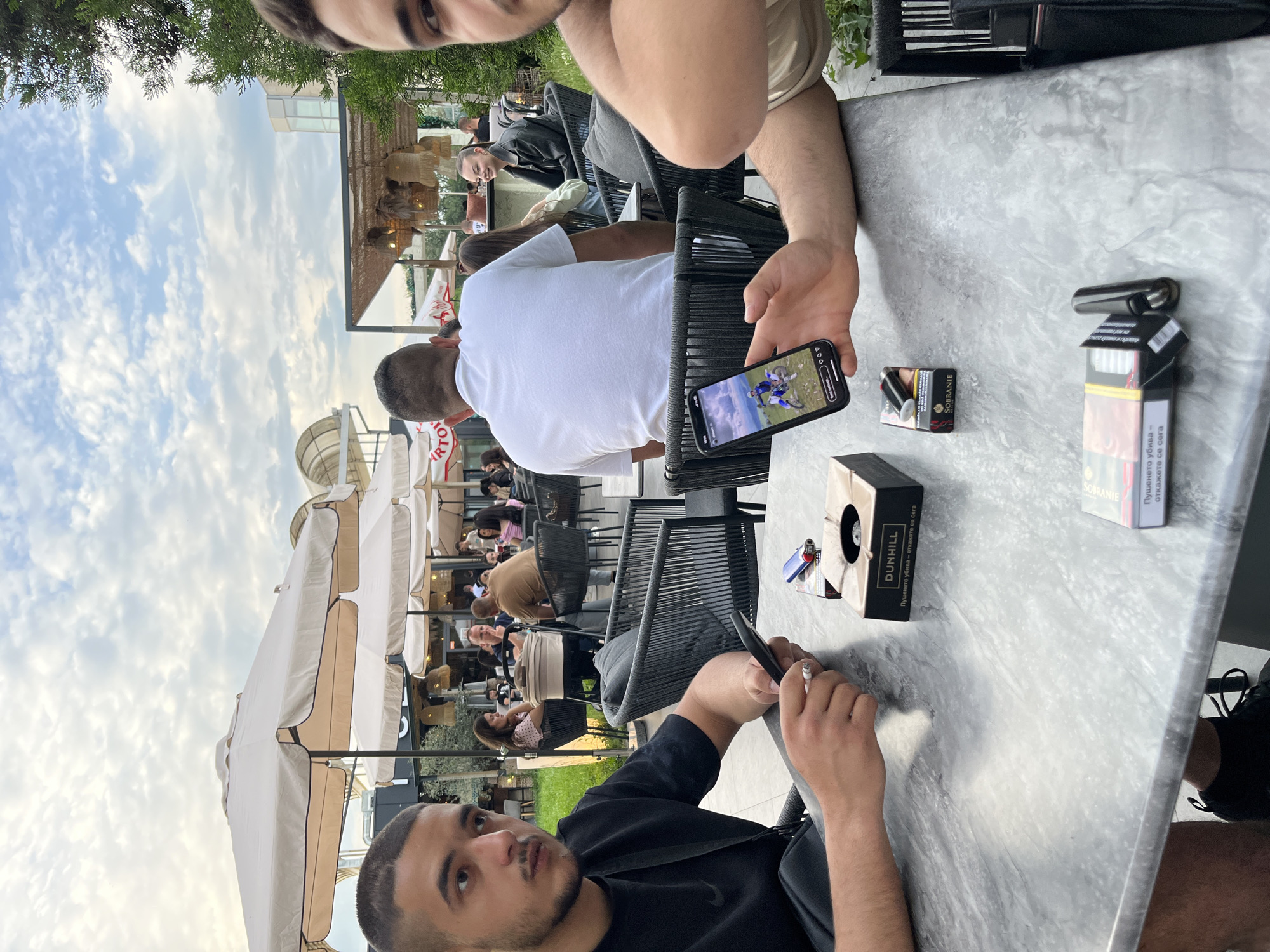 The image shows a group of people seated at tables in an outdoor dining area, likely a restaurant or cafe. The location is indicated by the presence of Bulgarian text on cigarette packaging, indicating it's likely in Bulgaria. Given the urban setting and the presence of identifiable branding, it can be surmised that it is in Plovdiv. Two men are prominently positioned in the foreground. One man is holding a smartphone displaying an image of motocross riders. On the table are a packet of Dunhill cigarettes, a pack of Sobranie cigarettes, and a cigarette lighter. Other people are seated at tables in the background under large parasols, indicating an outdoor seating arrangement. The overall scene suggests a relaxed social setting with people enjoying food, drinks, and conversation in an outdoor urban environment.