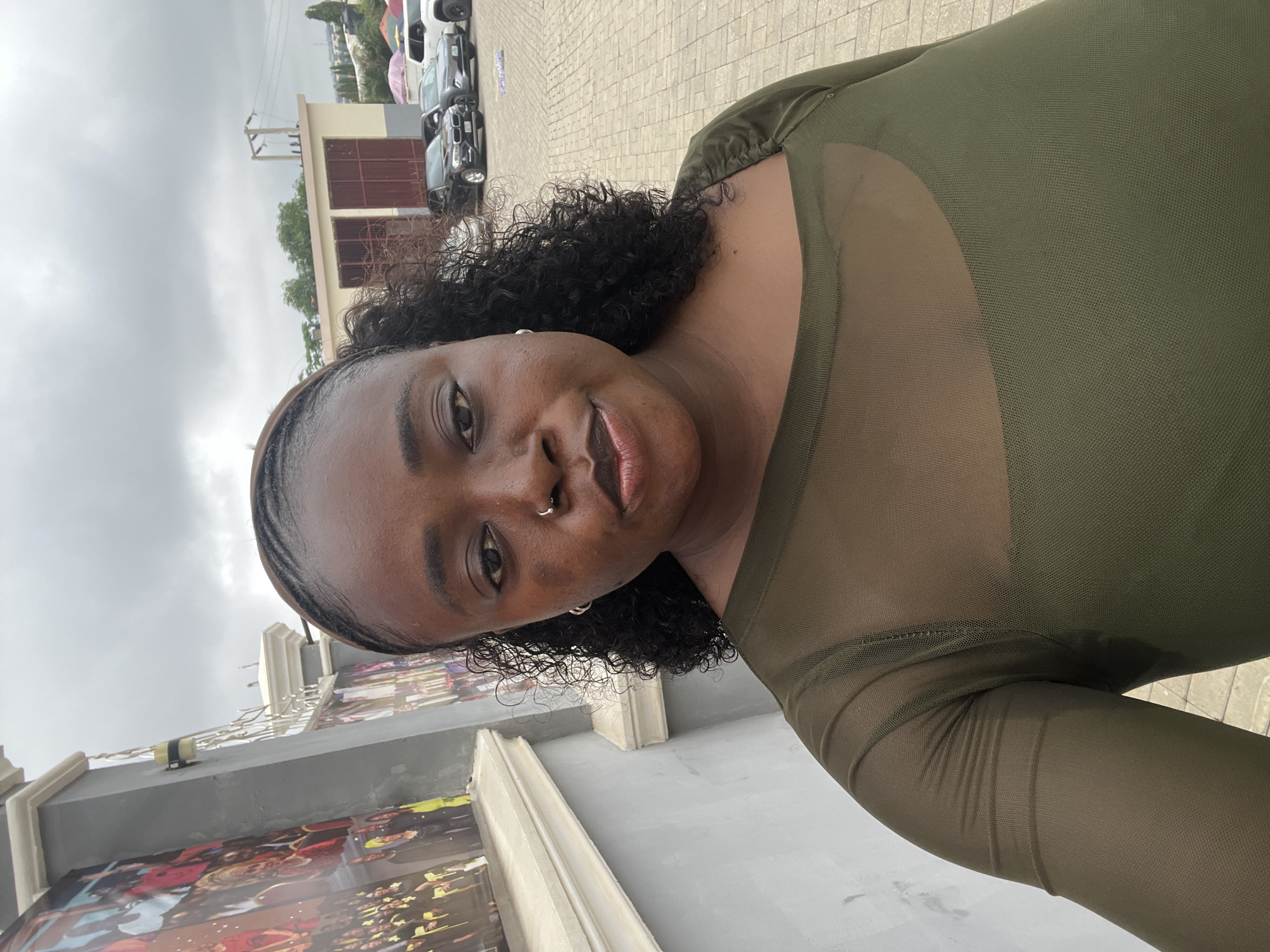 The image is a medium shot of a woman posing for a selfie outdoors. She has dark skin, curly black hair pulled back with a dark headband, and is wearing a sheer olive green long-sleeved top with a square neckline. She has a nose ring and small hoop earrings. The background shows a paved area with several cars parked, including a black BMW and a white SUV. Behind the cars, there's a beige building with brown doors and a power line tower. To the left of the woman, a grey wall with a textured white ledge is visible, adorned with large posters depicting groups of people in formal attire. The sky is overcast and cloudy. The scene appears to be in an open area, possibly a parking lot or event space, within the city of Abuja, Nigeria.