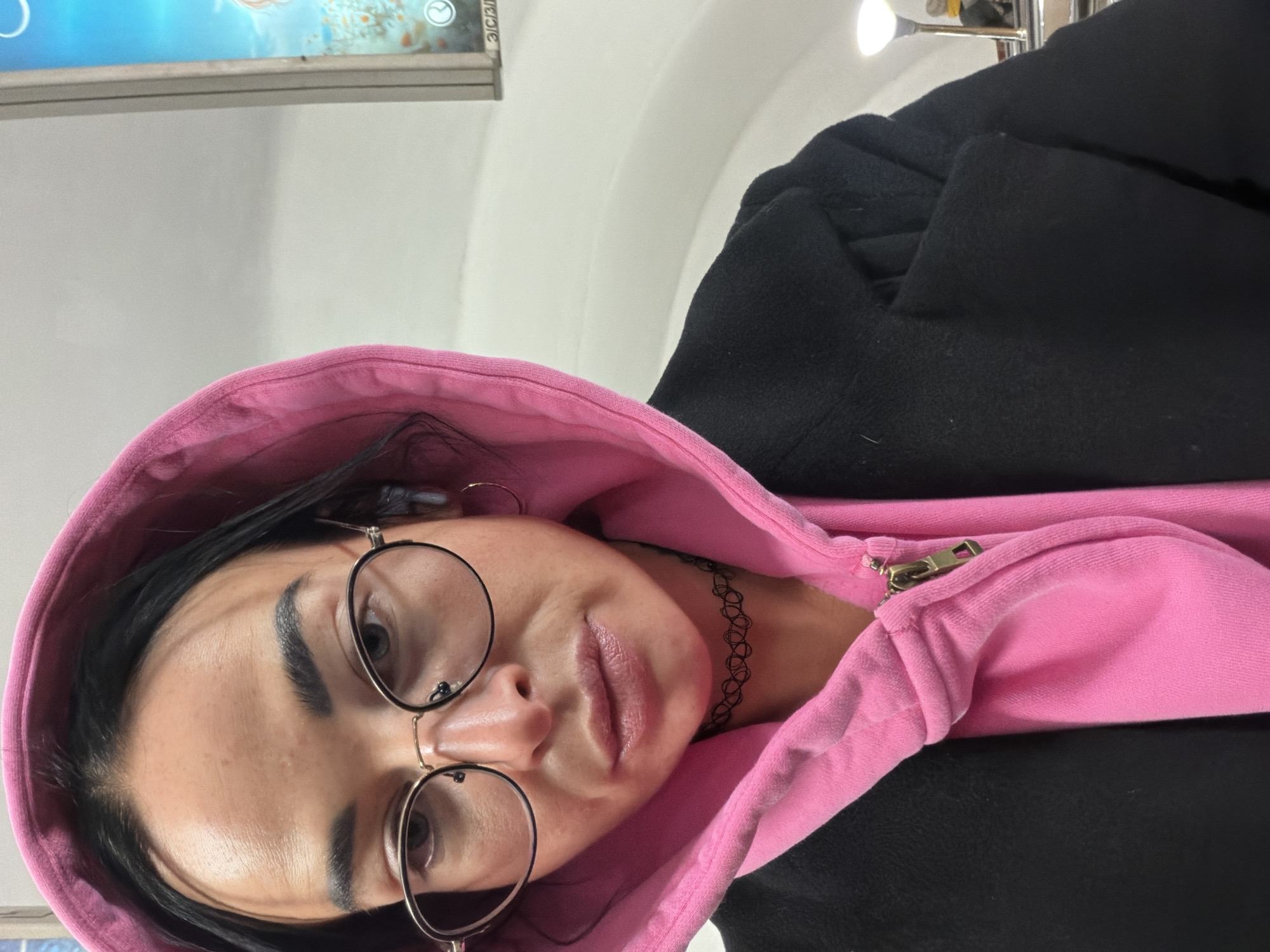 An overhead, close-up shot captures a person, likely a young woman, looking up towards the camera. The image appears to be a selfie or a photo taken from a high angle, set indoors in Saint Petersburg, Russia.
The subject has dark hair, styled to frame her face, and prominent, well-defined dark eyebrows. She wears round, gold-rimmed glasses that sit on her nose. Her lips are full and have a slight pout, contributing to a neutral or somewhat pensive expression. Around her neck, she wears a black, intricate choker necklace.
Her attire consists of a bright pink hooded sweatshirt or jacket, with the hood pulled up around her head. A brass-colored zipper is visible on the front of this pink garment. Over the pink layer, she wears a dark, possibly black, heavy coat or jacket, parts of which are visible on her shoulders and arms.
The background is predominantly a light-colored, possibly white, wall or ceiling. In the upper left corner, a portion of a digital display screen is visible, showing abstract or aquatic blue and white graphics and a small circular logo. Some faint text on the screen, partially obscured, appears to include Russian words like "ЭЛЕКТРОННЫЙ" (ELECTRONIC) and "3D," aligning with the Saint Petersburg context. Bright artificial light sources are visible in the upper right background. The scene suggests an urban indoor environment, possibly a public space.