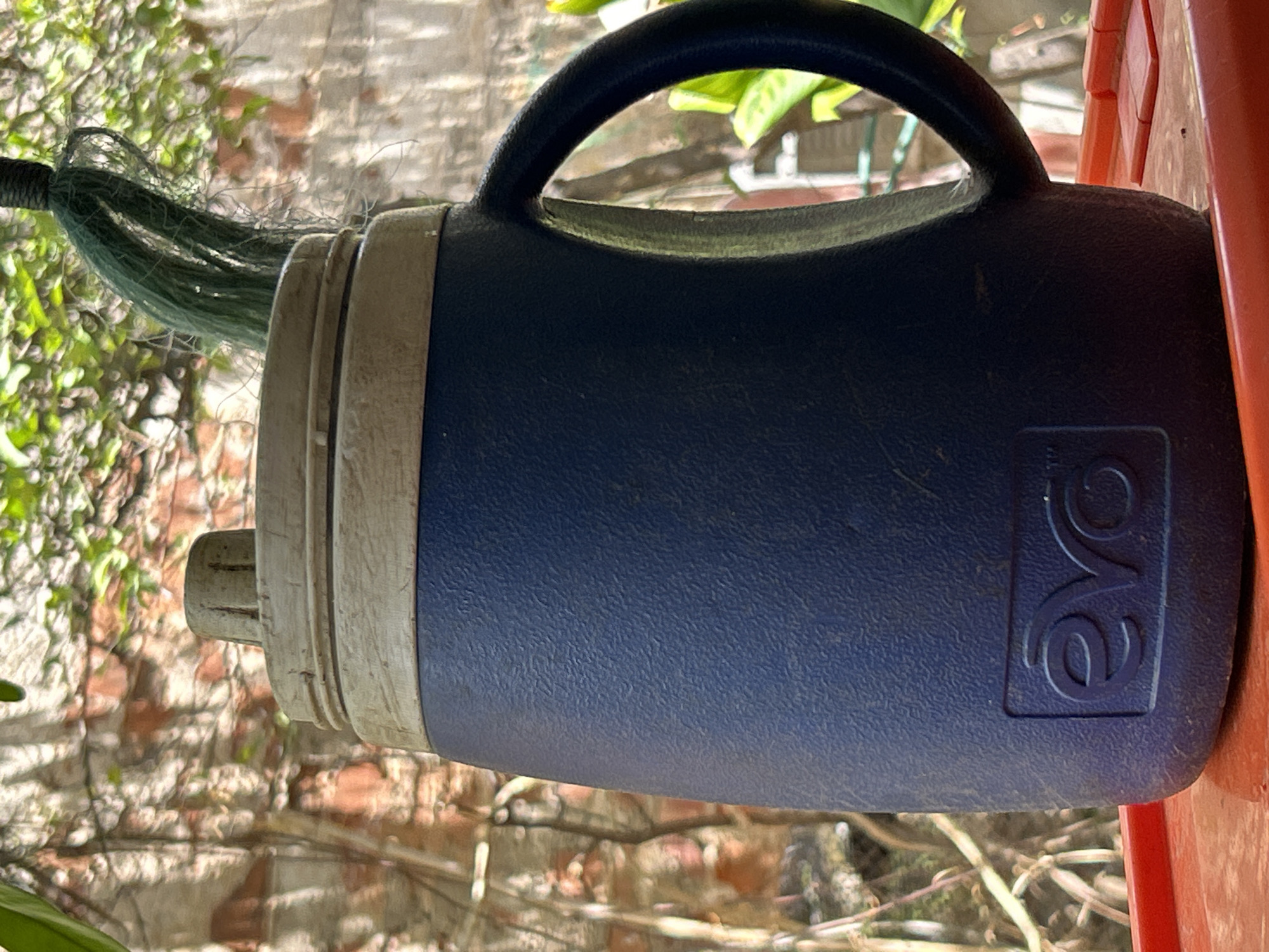 An outdoor, close-up shot captures a dark blue insulated pitcher with a textured surface. A beige lid with a beige cap is at the top, and a dark handle is attached to the side. A green string is hanging from the top of the pitcher. The pitcher is resting on a red surface. The background consists of a brick wall and foliage. The scene is located in Santa Ana, Venezuela.