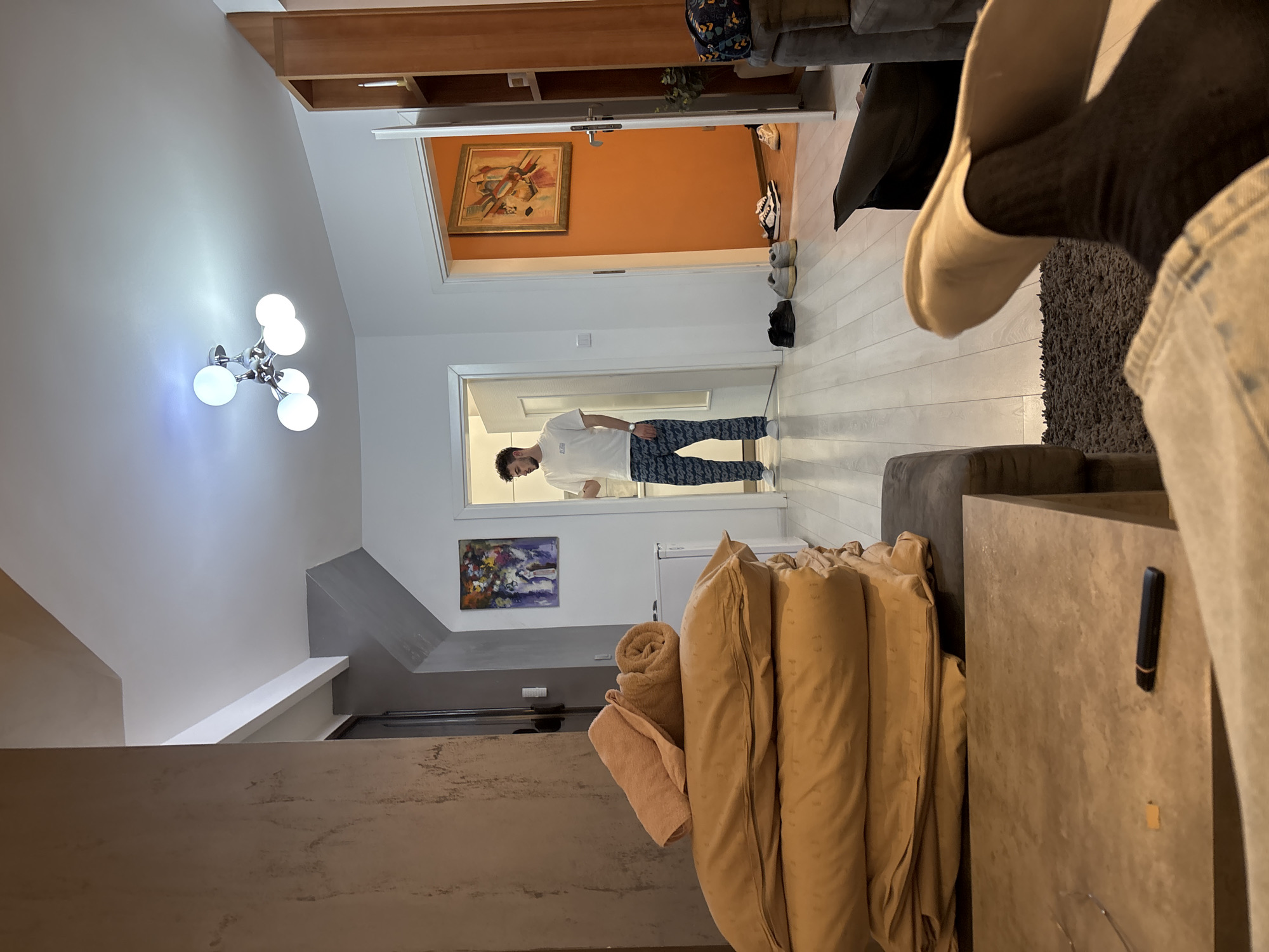 The image captures an indoor scene, likely a modern home in Plovdiv, Bulgaria, oriented horizontally. The perspective is slightly elevated, looking across a living area into a hallway.
In the center, a man with dark curly hair and a beard stands in a doorway, facing forward. He wears a white short-sleeved t-shirt and patterned blue and white pants. His posture suggests he is paused or observing.
The living space features light wooden flooring and white walls. A prominent modern chandelier with five spherical bulbs illuminates the left wall. To the right, a gray sofa or sectional is partially visible in the foreground, with another part forming a surface that holds a small black rectangular object. Stacked tan-colored folded textiles, possibly blankets or cushions, are neatly arranged on a low surface next to a white refrigerator. A rolled tan towel and a lighter-colored folded towel are also present.
Further into the background, the hallway reveals an accent wall painted a vibrant orange, adorned with a framed abstract painting in warm tones. Below this, on a white wall, is another framed painting with cooler, abstract colors. Several pairs of shoes, including sneakers and slip-ons, are arranged on the floor in the hallway. A close-up view shows a foot wearing dark socks and a light-colored slip-on shoe, positioned on a dark rug.
The overall atmosphere is well-lit, with a mix of artificial light from the chandelier and general brightness suggesting daytime. There is no visible text in the image.