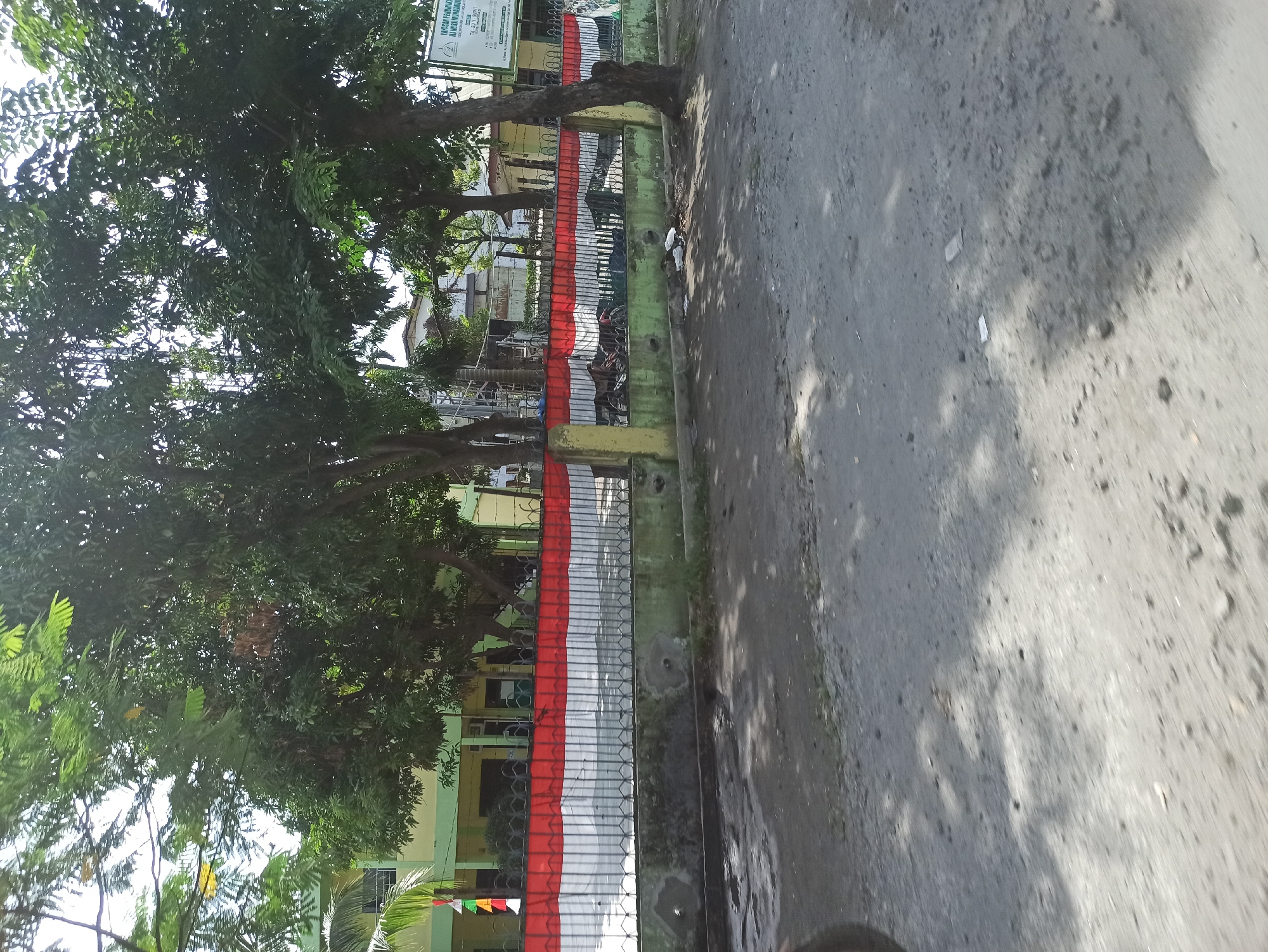 The image shows a street-level view of a building, likely a school, in Manunggal, Indonesia. The building is painted in light yellow with green trim. A fence runs along the front of the property, decorated with the Indonesian flag, red and white stripes. Trees with dense green foliage are situated in front of and around the building, casting dappled shadows on the ground. The street in the foreground is unpaved and appears to be made of dirt and gravel, with some scattered debris and shadows from the trees. A sign is visible on the right side of the building, though its text is not entirely legible. The overall scene depicts a typical day in a residential or educational area.