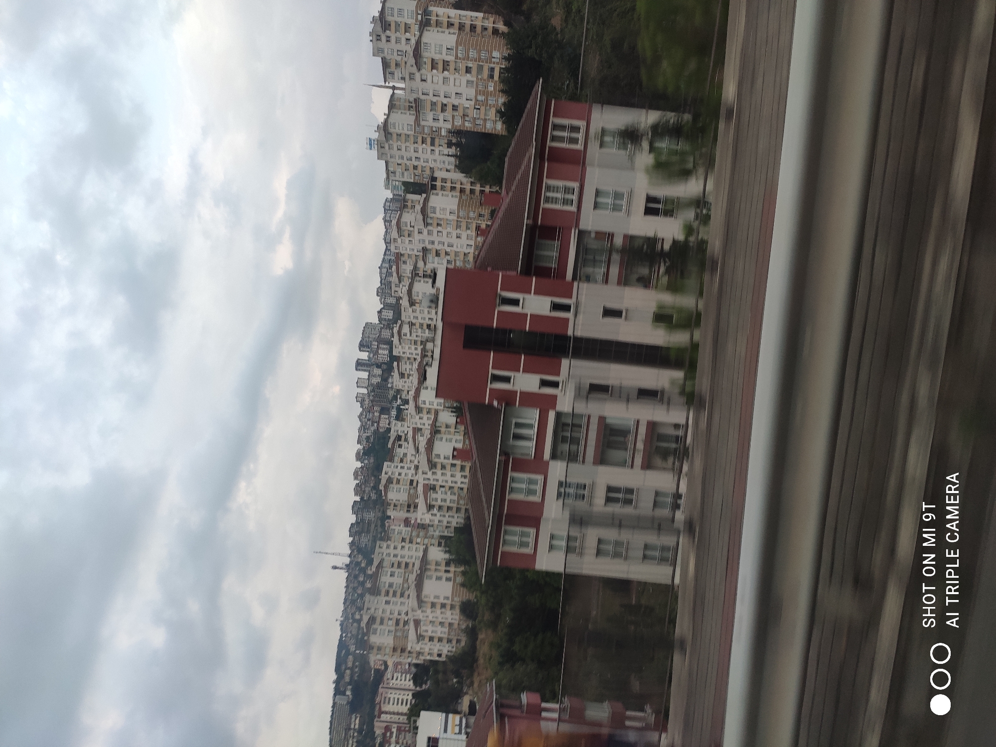 The image depicts a panoramic view of a densely populated urban area in Trabzon, Turkey, with a focus on residential buildings. The scene is dominated by a multitude of apartment complexes, primarily in shades of white, beige, and light brown, cascading up a hillside under an overcast sky. The architecture suggests a modern urban development, with numerous balconies and windows visible on the buildings.
In the foreground, a red and white apartment building stands out. It is a multi-story structure with a dark red roof and prominent white trim around the windows and balconies. Adjacent to this building, another structure with a red facade is partially visible. A metal guardrail runs horizontally across the bottom portion of the image, suggesting the photograph was taken from a moving vehicle, likely on a highway or elevated roadway. The blurred motion further supports this observation.
The environment appears to be a hillside overlooking the city, with lush green trees interspersed among the buildings, particularly on the lower slopes. The sky is filled with grey clouds, indicating a possibly cool or rainy day. The overall impression is one of a sprawling urban landscape characterized by consistent architectural styles and a sense of density. No individuals are visible in the frame. The text at the bottom right corner, "SHOT ON MI 9T AI TRIPLE CAMERA," is a watermark from the device used to capture the image.