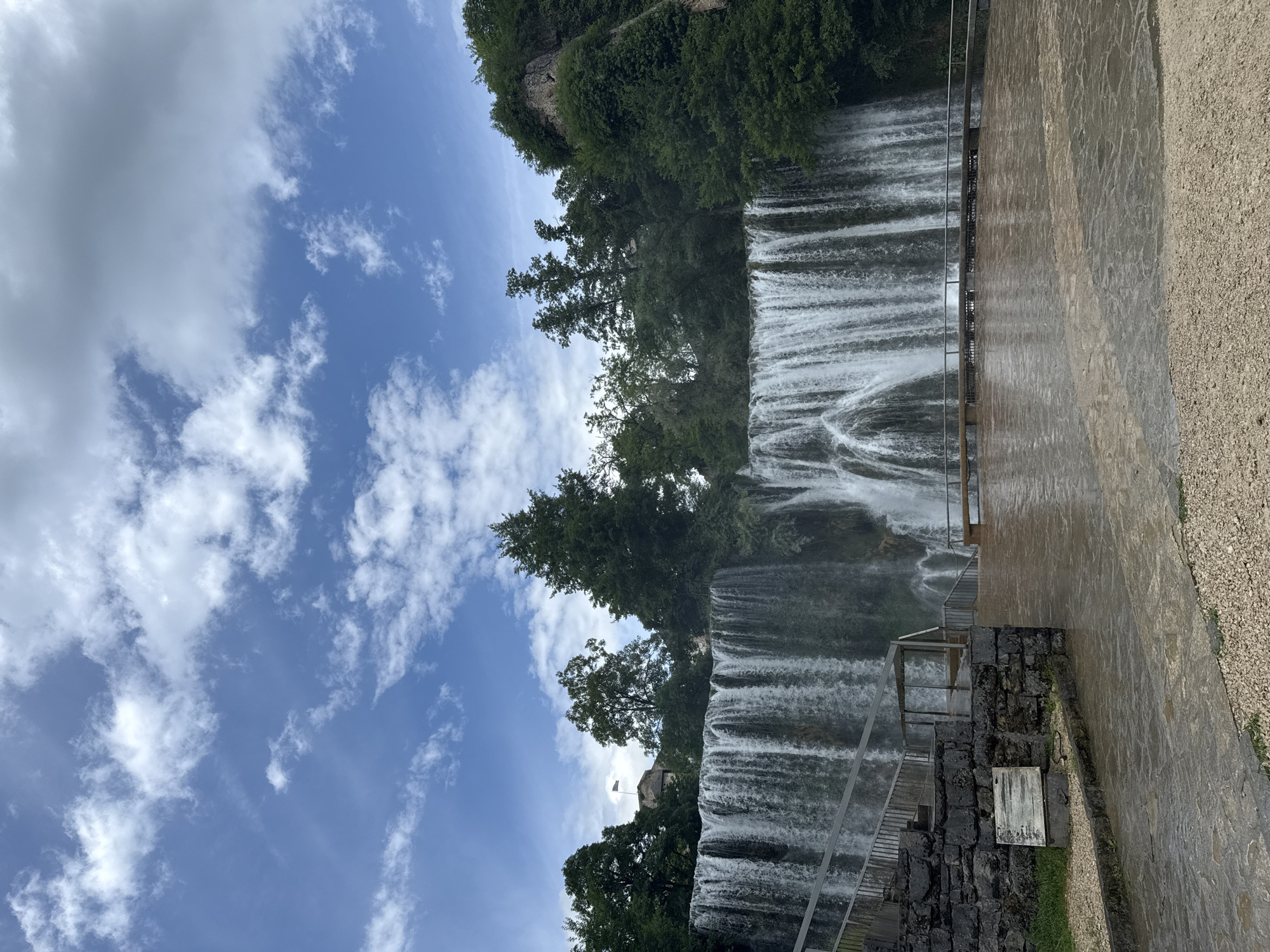 This full-shot image depicts the Pliva Waterfall in Jajce, Bosnia and Herzegovina, under a sky with scattered white and light gray clouds. The wide waterfall spans the mid-ground, presenting multiple cascades of white water descending into a visible wet, paved area below. Above the waterfall, a dense line of green trees extends across the scene. On the left side, partially obscured by trees, a stone building with a dark roof is visible, topped by a flag flying from a pole. To the right, a rocky cliff face, partially covered in vegetation, rises above the waterfall. In the lower-left corner, a dry-stacked stone wall supports a metal railing system with steps ascending. A rectangular, faded white sign is affixed to this stone wall. The immediate foreground features a wide, wet, dark gray paved area composed of irregular flagstones, bordered by light brown gravel at the bottom. A metal railing system delineates the edge of this paved area closer to the waterfall, and a stone bench is positioned on the far right of the paved area. No people are present in the scene.