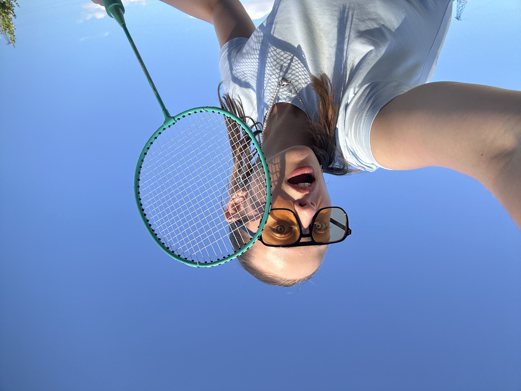 In an outdoor, low-angle, wide-shot, a young Caucasian woman with brown hair pulled back in a ponytail, wearing a light blue t-shirt, a silver heart-shaped pendant, and large, square, amber-tinted sunglasses, is playing badminton against a clear blue sky. She is holding a teal badminton racket in her right hand, which is extended towards the camera, and her mouth is open in a surprised or excited expression. The background is entirely the sky, with a hint of a distant industrial structure on the lower left and some foliage on the lower right. The lighting indicates it is daytime and sunny. The image appears to be a selfie taken in Khimki, Russia.