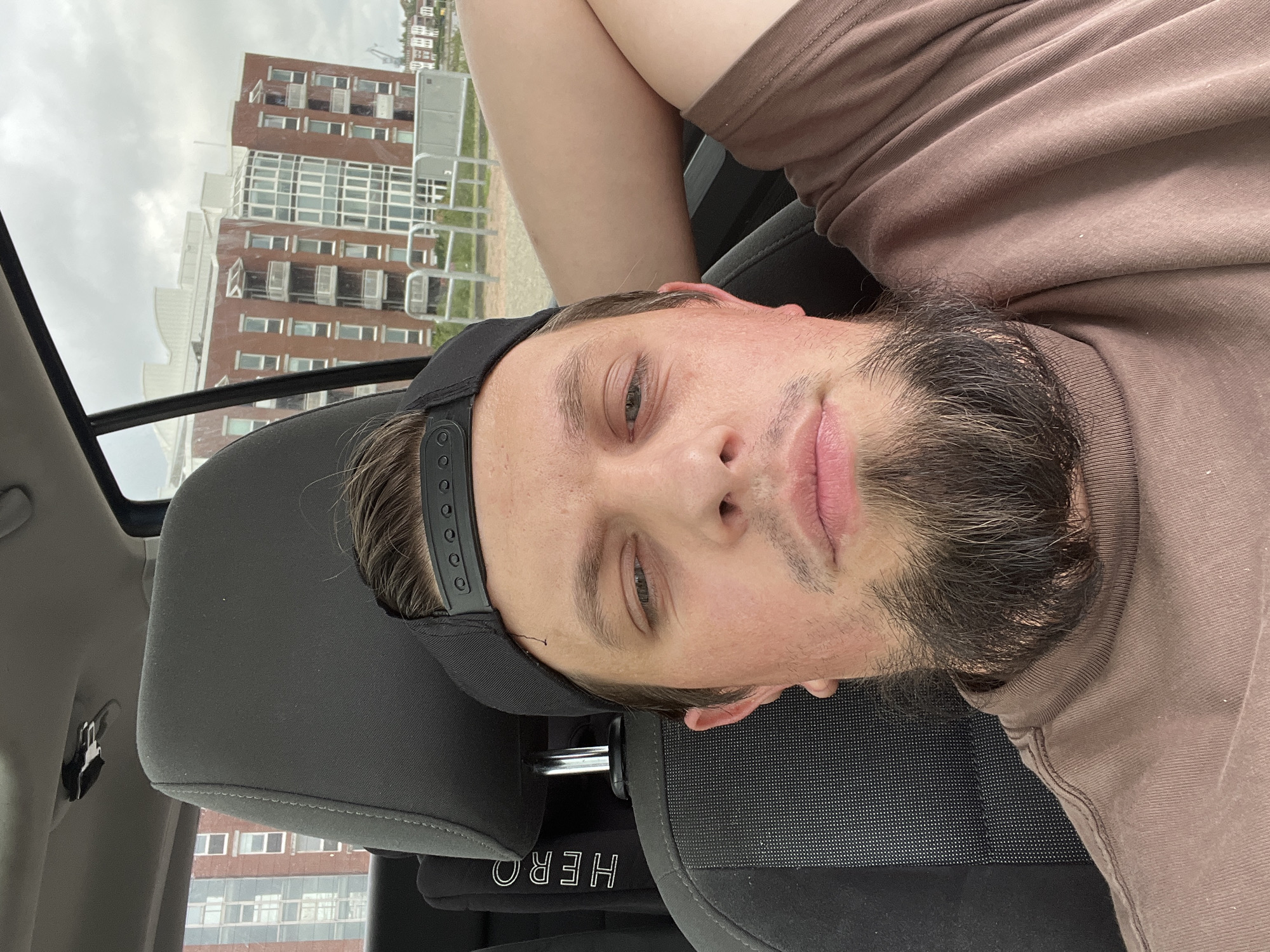 A man wearing a brown t-shirt and a black baseball cap is in the driver's seat of a car. He has a beard and is resting his arm on the headrest. In the background, a brick building with multiple windows and balconies is visible. This building is located in the port area of Wilhelmshaven, Germany, specifically near the new building complex called "Speicherinsel".