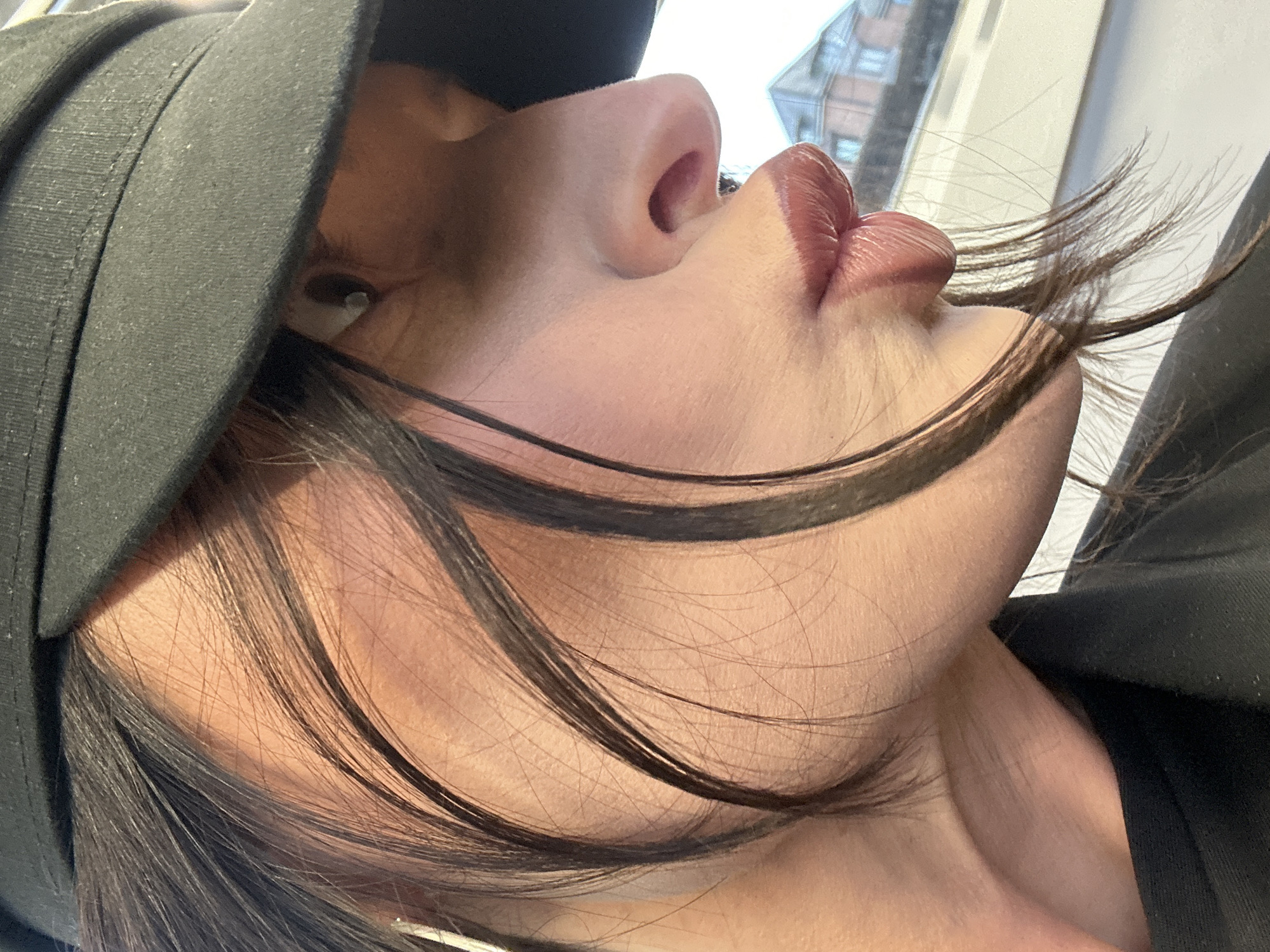 The image displays a close-up, profile view of an individual, likely female, posing with pursed lips in a pout. The subject wears a dark, possibly black, fabric cap that partially covers the forehead and eyes, revealing a visible white earbud in the right ear. Dark brown hair, with several strands framing the face, is present. The individual's fair skin shows natural texture. Lips are full, glossy, and coated in a reddish-brown color. A black garment is visible on the upper body. The background is out of focus, showing a brightly lit area and the upper section of a light-colored building featuring dark windows and a grey mansard roof. The scene is situated in the city of Tunbridge Wells, United Kingdom.