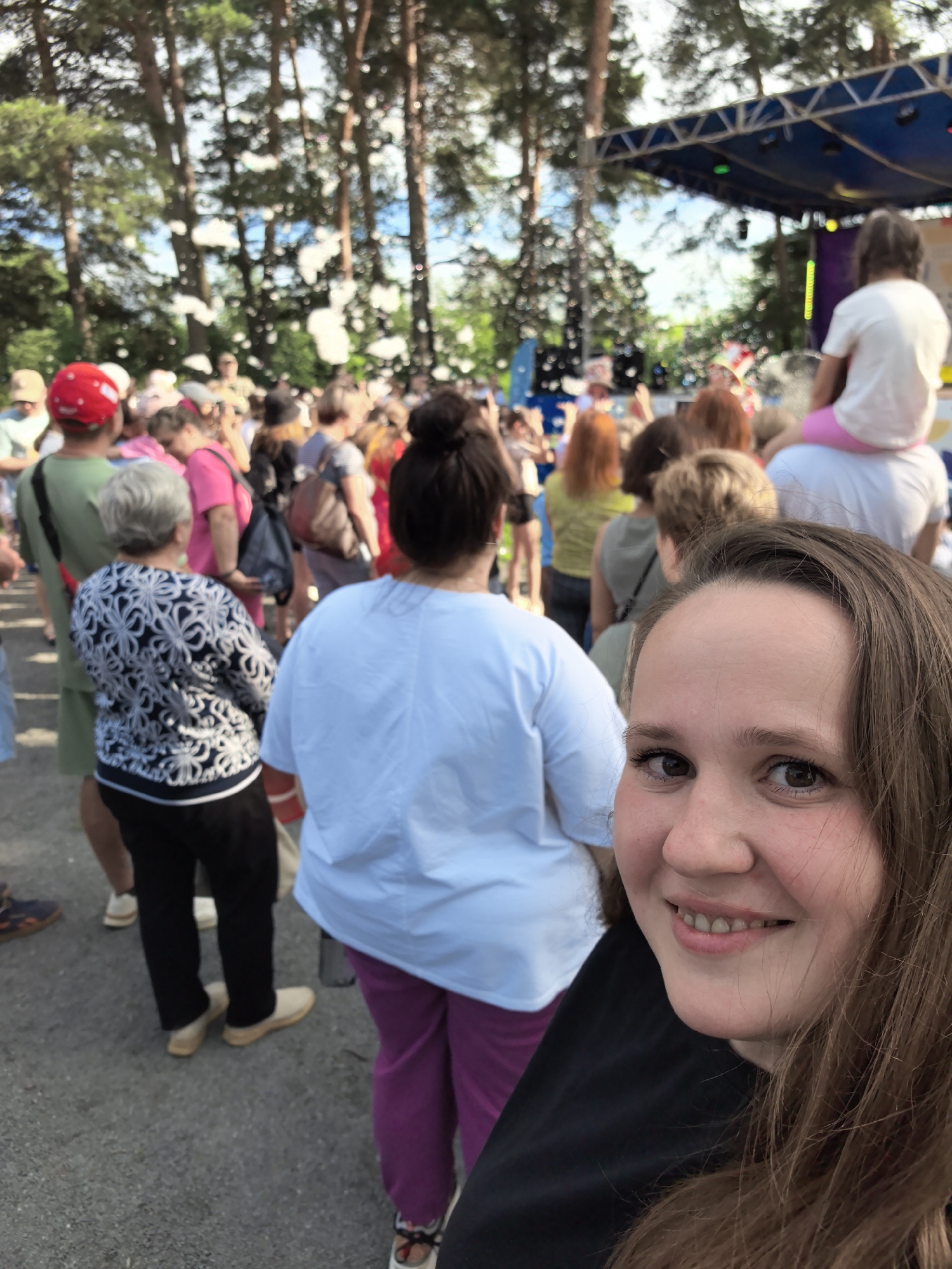 A medium shot captures a group of people at an outdoor event, with a young woman's face prominently in the foreground, smiling at the camera. She has long brown hair and is wearing a black shirt. Behind her, other attendees are visible, some with their backs to the camera, dressed in casual clothing. The background suggests a festival or concert setting with a stage and trees. The event appears to be taking place in the city of Gorny, Russia, as indicated by the surrounding pine trees and the general atmosphere of an outdoor gathering. Several people are wearing brightly colored clothes, and there are some blurry white elements in the air, possibly soap bubbles or confetti, adding to the festive ambiance. The ground is paved, and a crowd is gathered, indicating a public event.