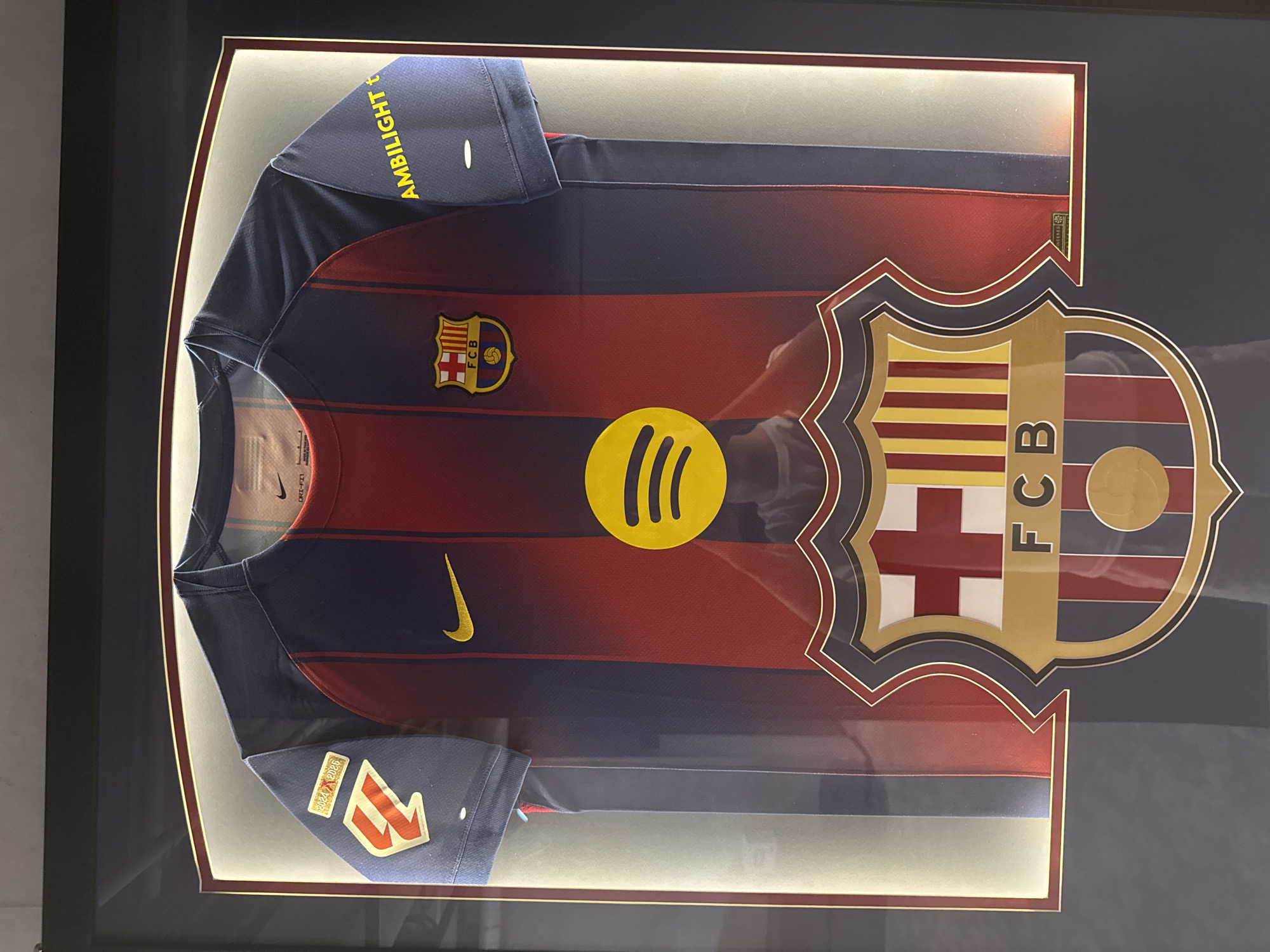 The image displays a framed FC Barcelona soccer jersey, likely a collectible, in a dimly lit setting. The jersey is predominantly red and navy blue, featuring the team's crest and a prominent yellow Spotify logo on the chest. The crest is a shield with diagonal red and yellow stripes, a white cross on a red background, and a blue background with a gold ball. The text "FCB" is visible within the crest. The jersey also has a yellow Nike swoosh on the chest. On the left sleeve, yellow text reads "AMBILIGHT t". Below the main jersey, on the bottom left sleeve, there are two patches: one reads "2024-2025" and the other has a stylized red and white design. The jersey is mounted within a frame, illuminated by internal lighting that creates a soft glow around it. The overall presentation suggests a display in a fan's home or a sports memorabilia shop. The background is dark, with hints of a reflective surface. No people are visible. The time of day and weather are not discernible. The location context of Kyiv, Ukraine is noted but not visually evident in the image itself.