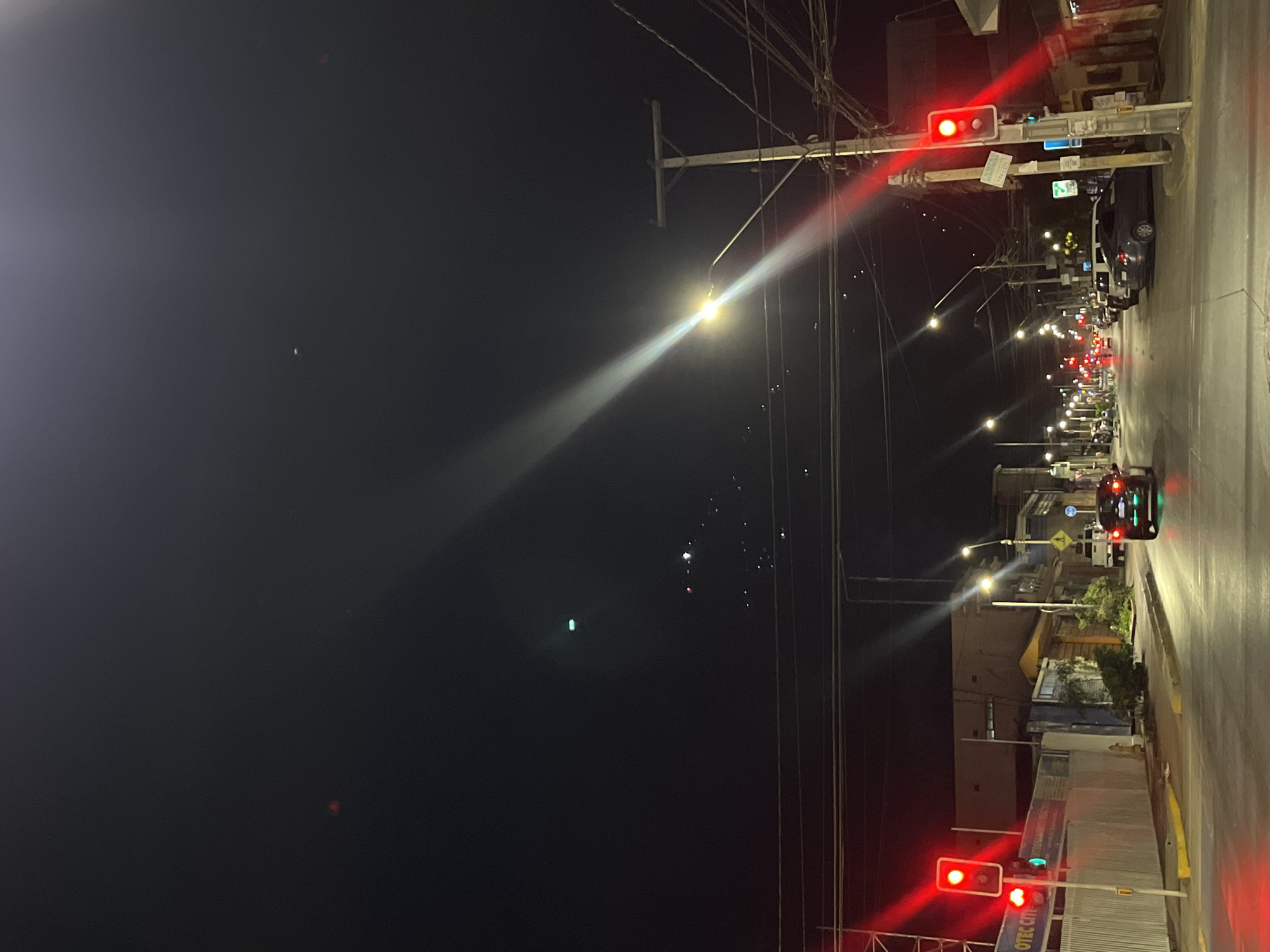 The image shows a nighttime street scene in a city, with an unknown location. The street is lined with streetlights and buildings. There are multiple cars. At both ends of the photo there is a traffic light which is red. Electrical wires are visible.