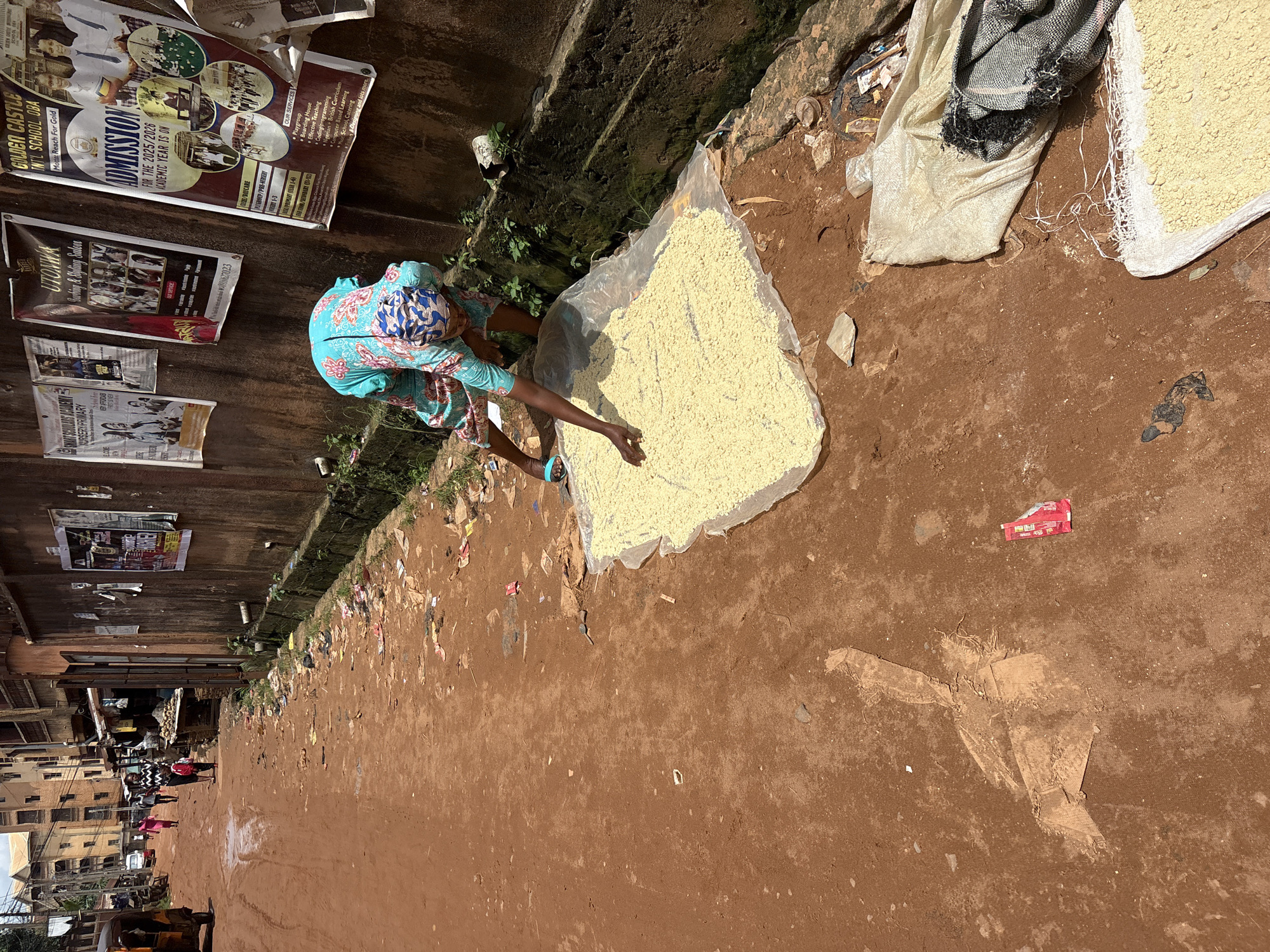 In the city of Nkpor, Nigeria, a woman in a blue and patterned headscarf and teal, patterned dress is crouched on a dirt road, sifting through a large pile of yellow granular material spread on a clear plastic sheet. The road is unpaved and appears muddy in places, with debris scattered along the sides. Behind the woman, a wall is covered with several posters advertising local businesses and educational institutions. To the left of the frame, a three-wheeled vehicle is parked. In the background, multi-story buildings line the street, suggesting an urban or semi-urban environment. The overall scene depicts an outdoor market or street-side activity, likely related to food preparation or drying, under a bright, overcast sky.
