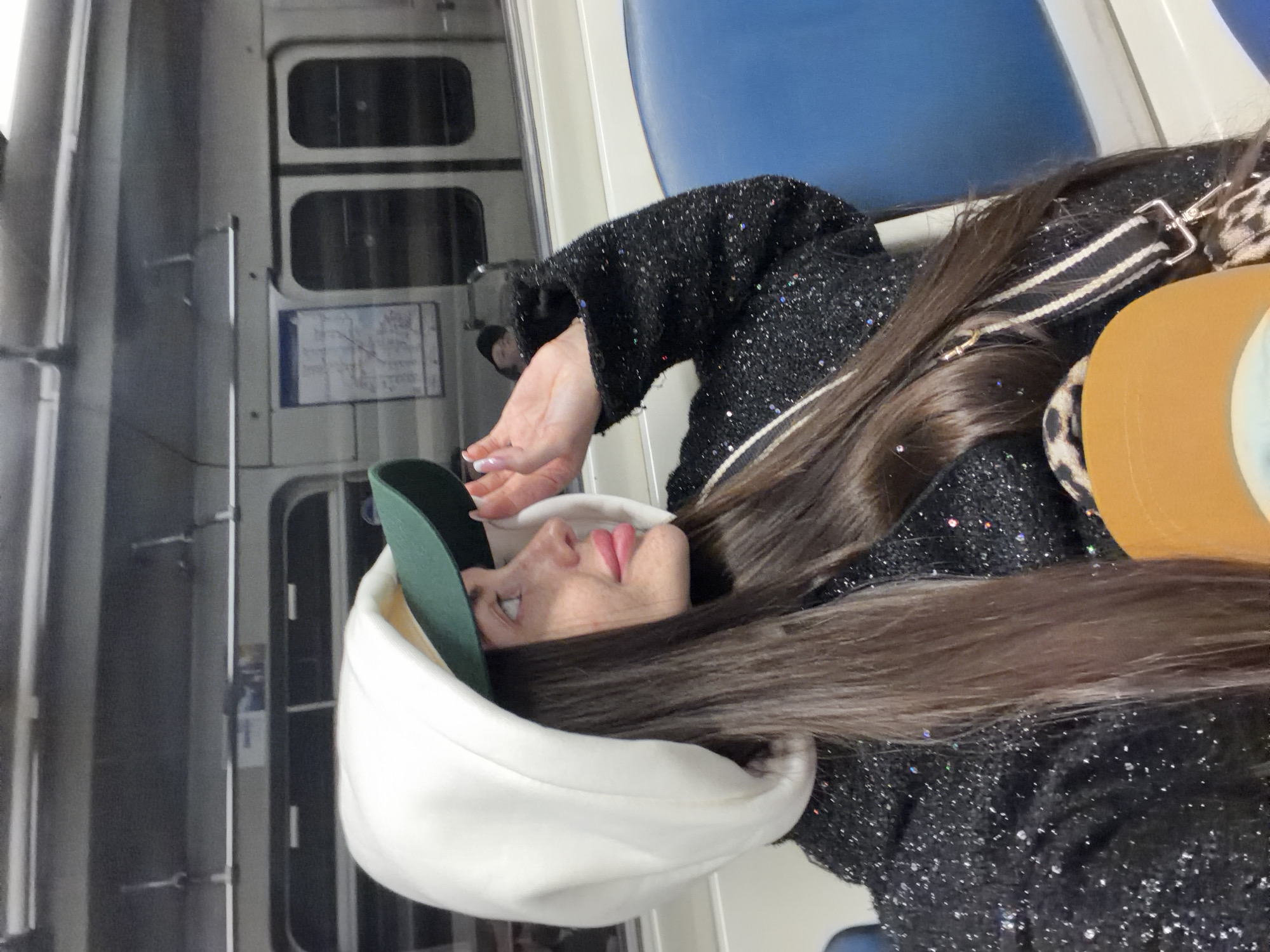 The image, rotated 90 degrees clockwise, depicts a woman inside a metro or train car, viewed from a side profile. She has long, straight dark brown hair that cascades over her shoulder. She is wearing a dark jacket or coat adorned with subtle sparkles. On her head, she wears a white, possibly hooded, garment, under which a dark green baseball cap is visible. Her complexion is fair, with noticeable freckles on her nose and cheeks, and her lips are a soft pink. Her right hand, showing manicured nails, is raised near her face. She carries a bag with a distinct black and white striped strap and a section with leopard print. An additional tan or orange object is also visible, possibly part of her bag or another item.
The setting is clearly the interior of a public transport vehicle. The background features metallic grey walls, typical train windows with rounded frames, and vertical metal grab handles. A portion of a blue upholstered seat is visible to the right. On one of the walls, there's a small, rectangular informational sign or map with indistinct text and a diagram. The lighting is artificial, suggesting an indoor environment. The woman's expression is neutral, with her eyes looking slightly upwards, indicating she might be observing something outside the frame or simply in a state of contemplation. No discernible text is legible on the visible sign. The scene is consistent with a metro environment in Saint Petersburg, Russia, as per the provided context.