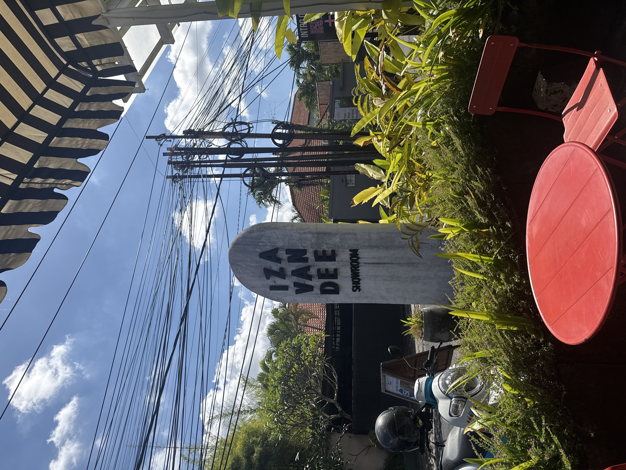 The image depicts an outdoor street scene in North Kuta, Indonesia, bathed in bright daylight under a clear blue sky with scattered white clouds. The perspective looks upwards, highlighting a dense network of black electrical wires crisscrossing against the sky, originating from utility poles in the mid-ground.
In the left foreground, a black and white striped awning extends, suggesting a covered outdoor seating area or shop entrance. To the right, a red outdoor table and part of two red chairs are visible, partially obscured by vibrant green plants and foliage that line the foreground and extend towards the middle of the frame, contributing to a lush, tropical setting.
The central subject is a tall, surfboard-shaped concrete sign prominently displaying "IZA VAN DEE SHOWROOM" vertically in black capital letters. Below this sign, a white and light blue Honda scooter is parked, with a black helmet resting on its seat and another helmet nearby. A wooden box on the scooter has partially visible text including "Breckar Special."
In the background, behind the sign and foliage, buildings with reddish-brown tiled roofs and dark walls are discernible, interspersed with palm trees and other tropical vegetation. A smaller sign in the background includes the text "INTRO +" and a QR code. The absence of people suggests a quiet moment in an otherwise bustling area. The scene portrays a typical commercial and residential environment in a tropical setting.