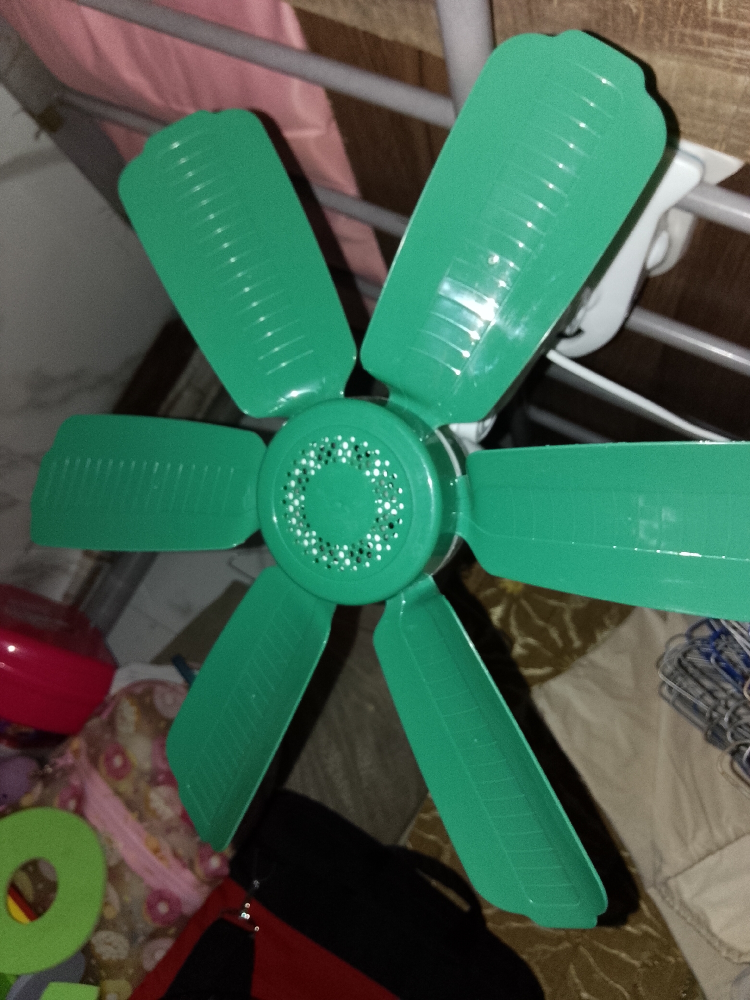 The image captures an indoor scene, likely a bedroom in Davao City, Philippines, focusing on a vibrant green electric fan. The fan features six broad, rectangular blades, each with subtle horizontal ridges, and a central circular housing with a pattern of small perforations. It is a clip-on type, affixed by a white clamp mechanism to a light grey metal frame, possibly part of a bed frame or shelving unit.
Behind the fan and the metal frame, a soft pink fabric, possibly a blanket or curtain, is visible. A dark wooden surface, likely a wall or piece of furniture, forms part of the background.
Below the fan, various items are present, suggesting a lived-in space. On the left, a portion of a red plastic bowl or container can be seen, next to a light green foam ring-shaped object. Further back and slightly out of focus, a pink object with white, donut-like patterns, possibly a child's bag or patterned textile, is visible. On the right, a stack of metal hangers rests on a light-colored fabric, which appears to be a bedsheet or blanket. A dark bag, potentially a backpack with red details, is visible at the bottom edge of the frame.
The lighting suggests an indoor setting, possibly illuminated by artificial light or experiencing dim daylight. No individuals are present, and no particular activity or event is taking place; the scene is a static depiction of a room's contents. There is no visible text on any of the objects. The fan's presence is consistent with the warm, tropical climate typical of Davao City.
