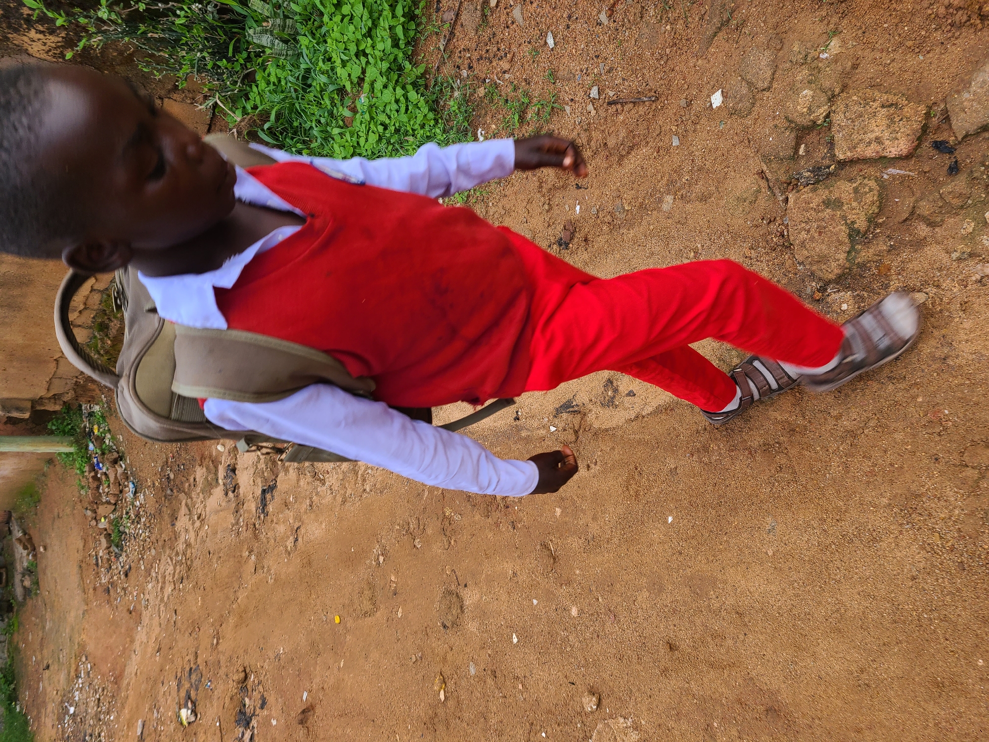 A young boy wearing a white long-sleeved shirt, a red V-neck vest, and red trousers walks on a dirt path. He carries a brown backpack with a tan strap over his left shoulder. His feet are clad in grey and white sandals with dark straps, and white socks. The path is uneven and covered in loose sand and small stones, with some larger rocks and debris scattered around. Lush green vegetation grows to the right of the path, adjacent to a textured, tan-colored wall or building. The scene appears to be outdoors in a somewhat rural or unpaved urban area, consistent with parts of Jos, Nigeria.