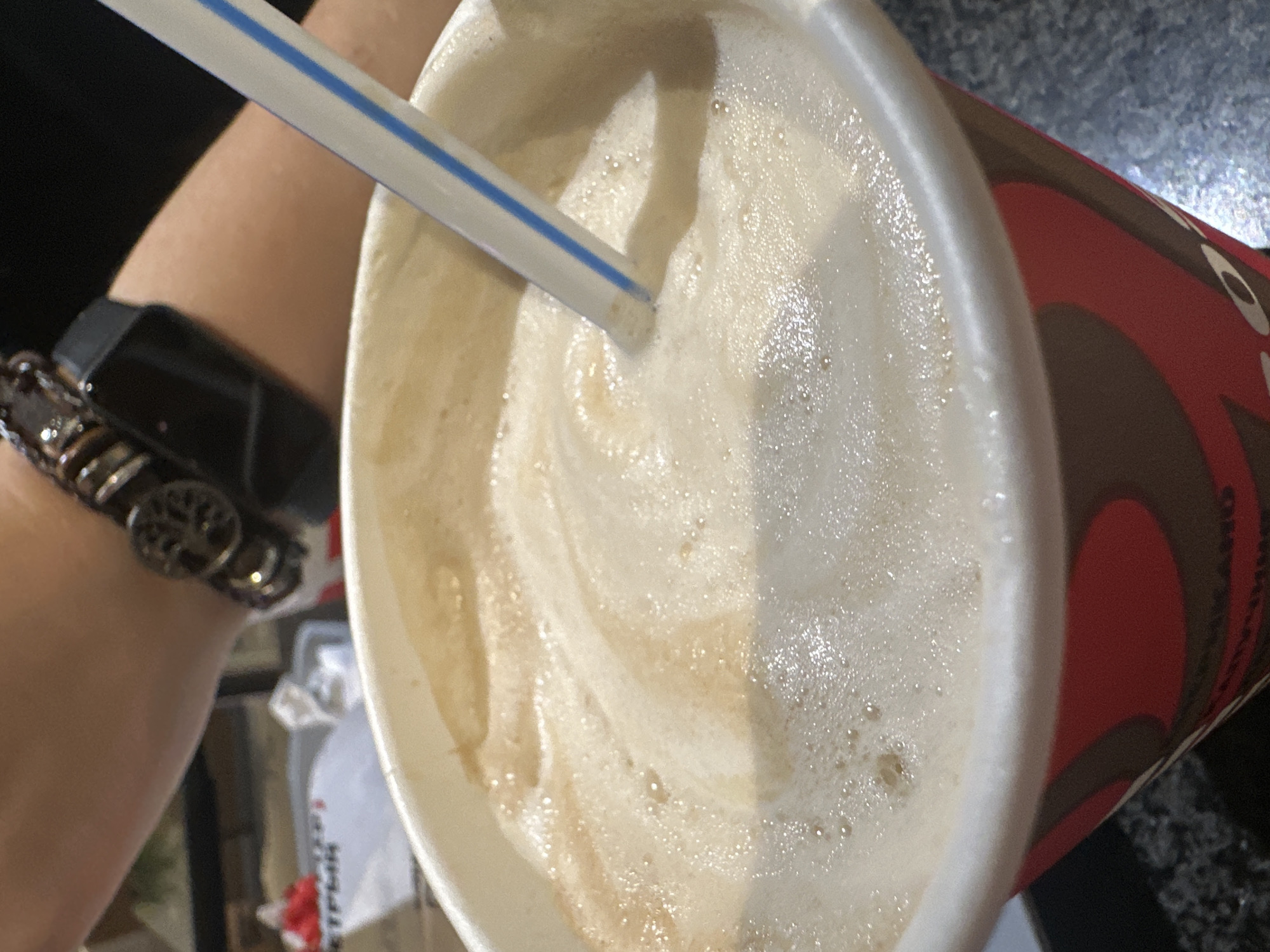 This is a close-up, eye-level shot of a disposable paper cup filled with a frothy, light brown beverage, likely coffee or a milkshake. A white and blue striped straw extends from the drink into the frame. In the background, a person's arm is visible, adorned with a dark bracelet featuring circular, metallic charms. The person is wearing a black smartwatch on their left wrist. The background also shows hints of a blurred environment, including a white paper bag with red lettering that might be from a fast-food establishment. The cup itself has a red and brown patterned design. The setting appears to be indoors, possibly a cafe or a casual dining area. There is no specific indication of the city of Stavropol, Russia, within the image itself.