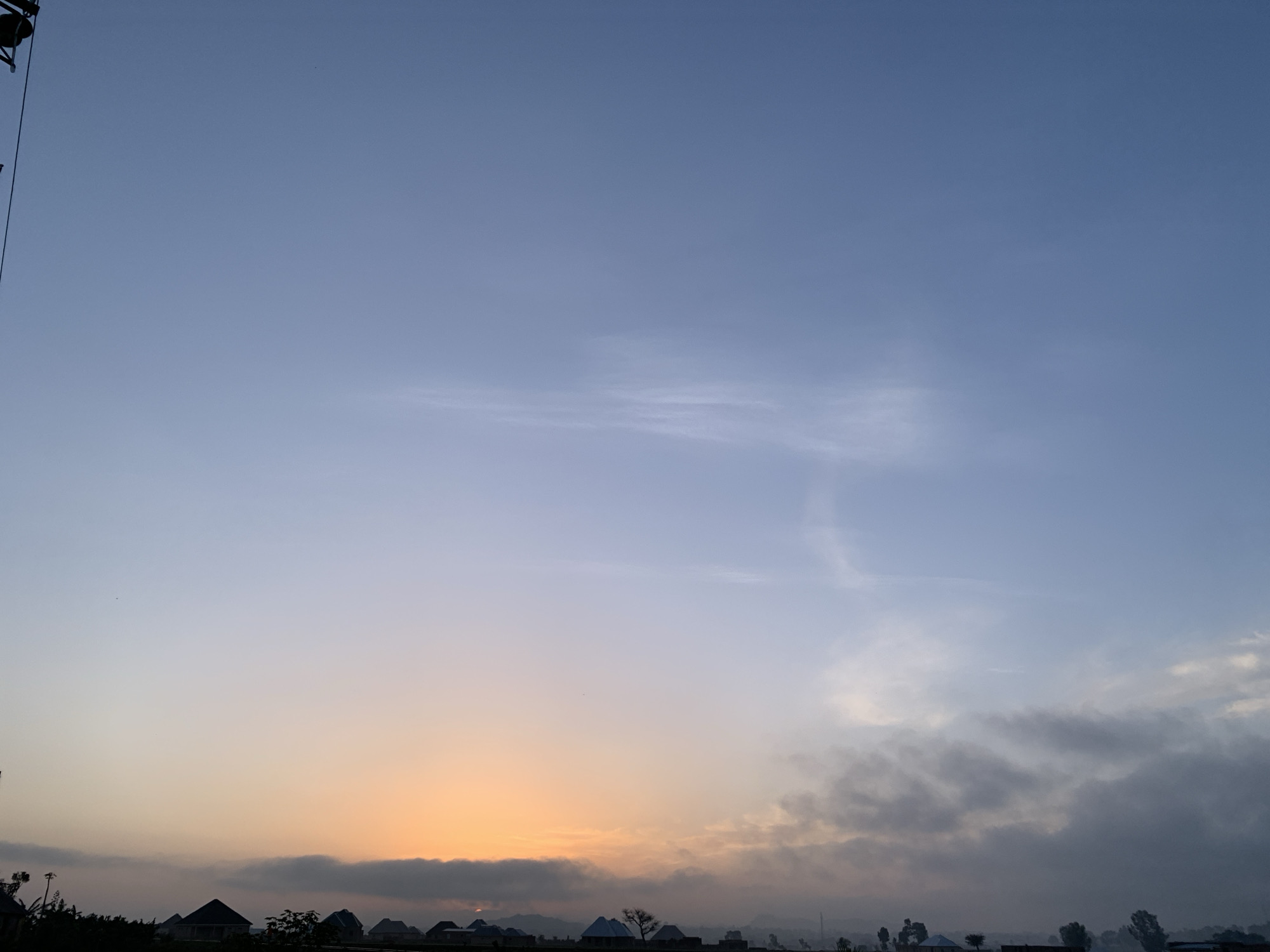 A twilight sky over Kuru Karama, Nigeria, displays a gradient of colors, transitioning from a pale orange glow near the horizon to a soft blue overhead. Wispy clouds are scattered across the sky, with denser, darker clouds accumulating towards the right side of the frame. Silhouetted against the brightening sky, the rooftops of several buildings, appearing to be homes, are visible along the horizon. The structures have distinct conical or pyramidal roofs, and some are adorned with intricate patterns. Trees and other vegetation form a dark line along the base of the scene, further obscuring the ground. The overall atmosphere suggests the early morning or late evening, with a tranquil and somewhat serene ambiance.