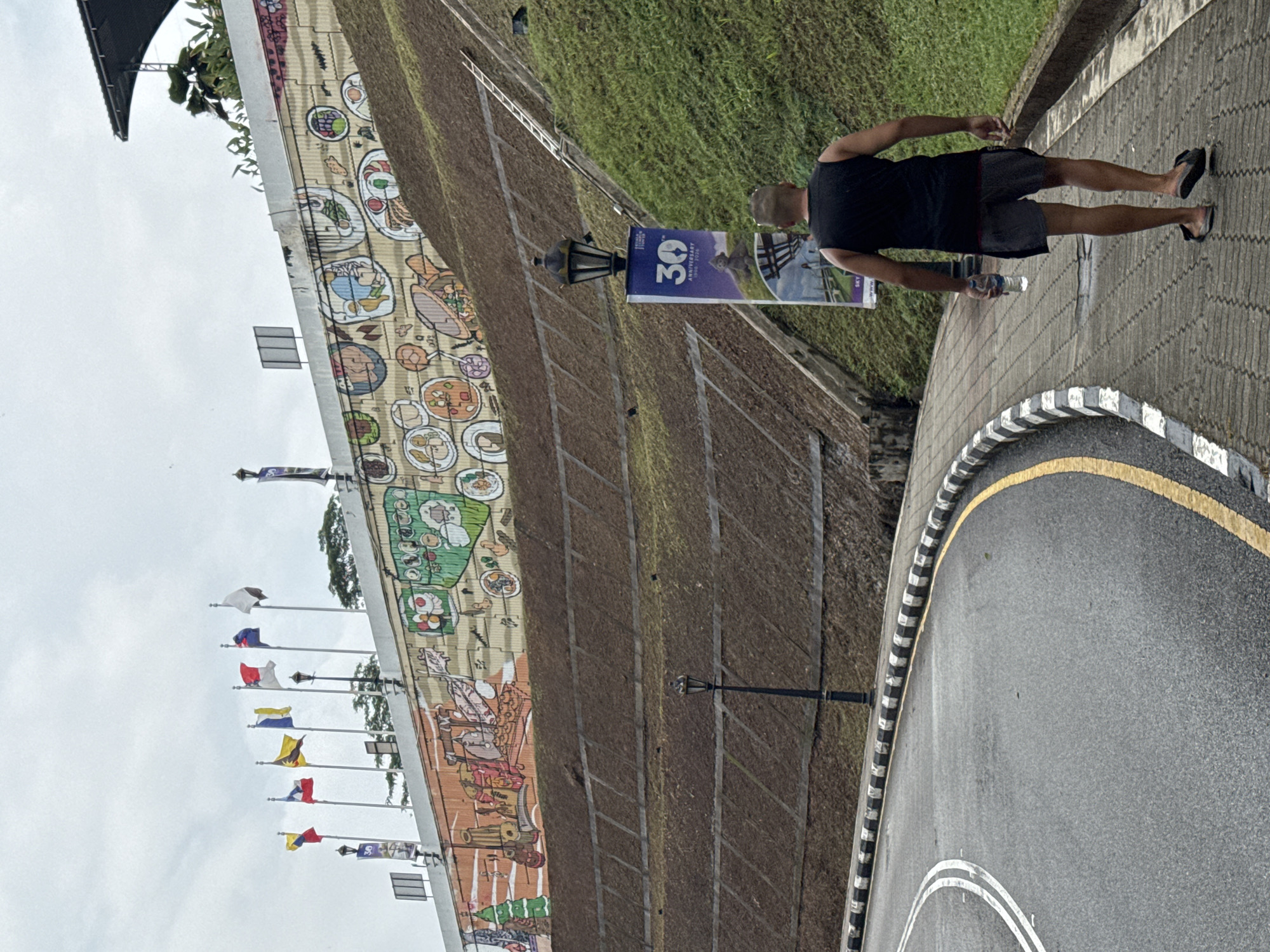 A medium-angle shot depicts a man walking on a paved road with a black-and-white striped curb in Kuala Lumpur, Malaysia. He is wearing a black tank top, dark shorts, and flip-flops, and he is holding a water bottle. To his left, a grassy slope leads to a tall retaining wall that is painted with a mural depicting various food dishes. Flagpoles displaying different flags are lined up along the top of the wall. A sign with "30th Anniversary" on it is positioned next to a street lamp along the wall. A road curves in the foreground. The sky is overcast.