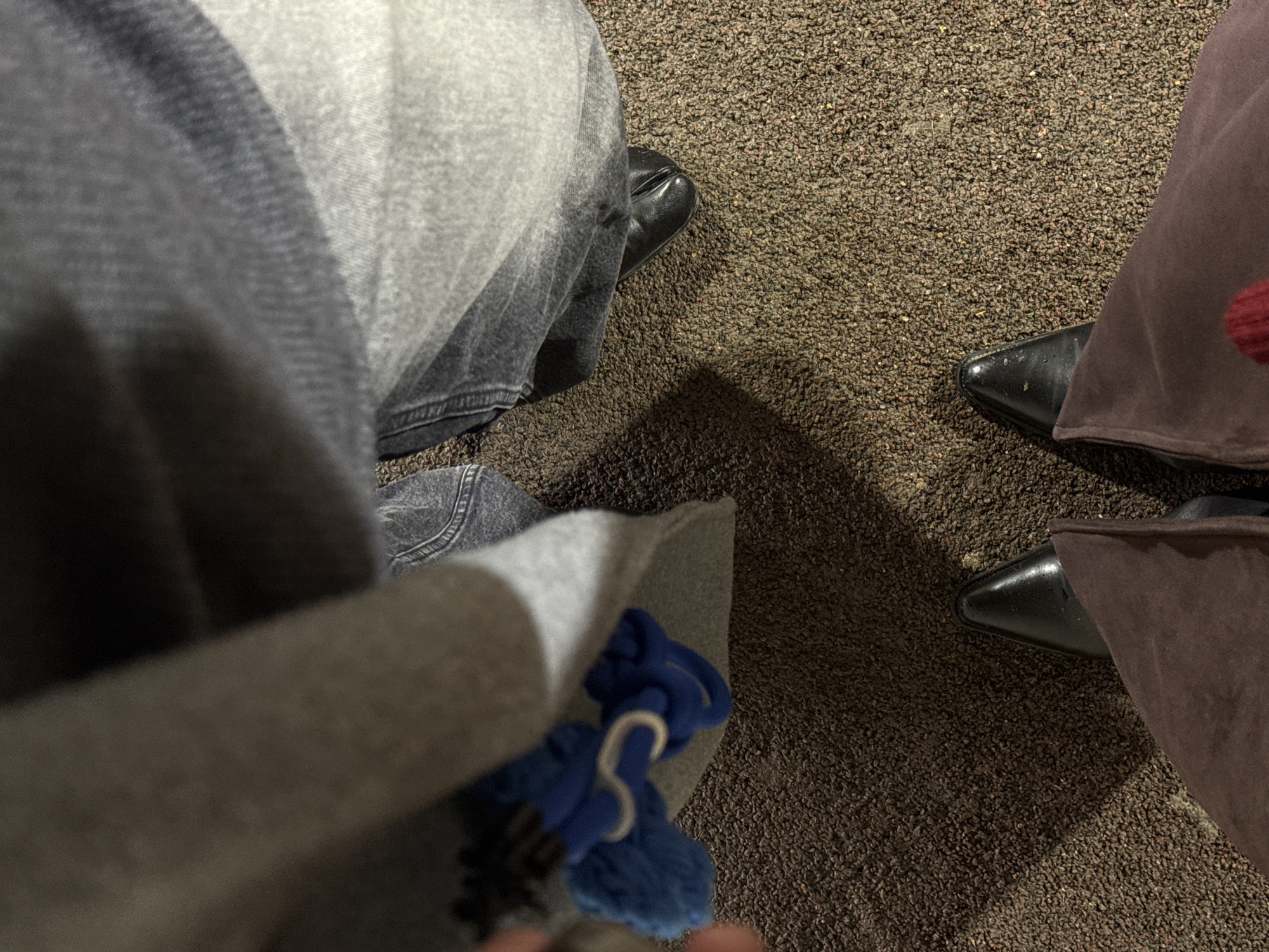 The image depicts a close-up view of two people sitting side-by-side, likely indoors given the carpet. The person on the left is wearing blue jeans and black shoes. In the foreground is a grey jacket or sweater with a blue object that has white ties/strings. The person on the right is wearing what appears to be brown pants and black pointed shoes. The location in the city of Perm, Russia is not specified.