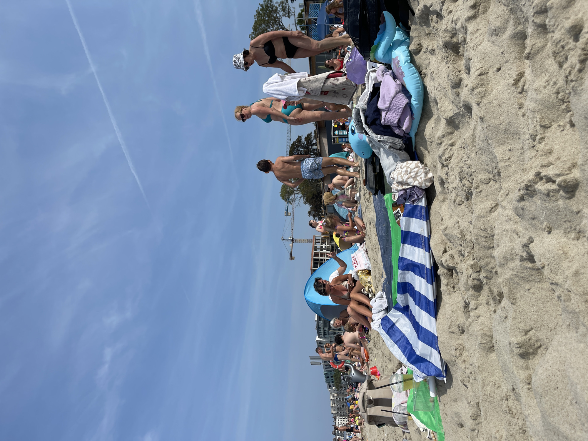 The image depicts a crowded beach scene with numerous people enjoying a sunny day. In the foreground, a pile of beach belongings, including a striped beach umbrella, towels, clothing, and inflatable toys, rests on the sand. Several individuals are seated or standing on the beach, some in swimwear, while others are engaged in activities such as talking or using their phones. In the mid-ground, more beachgoers are visible, along with a blue beach tent. In the background, buildings and a construction crane suggest the presence of a town or city. The sky is blue with some scattered clouds and contrails from airplanes. The overall impression is of a lively summer day at a popular seaside location. This image could be a beach in Zelenogradsk, Russia, which is known for its beaches.
