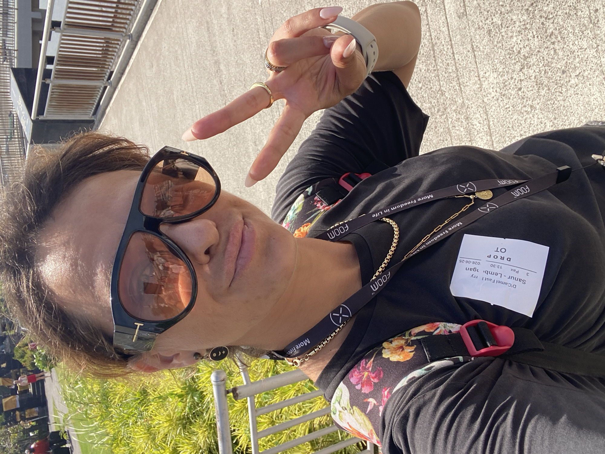 A woman, wearing a black t-shirt with a floral pattern, sunglasses, and a lanyard, takes a selfie outdoors. She is holding up two fingers in a peace sign with her right hand, which is adorned with rings and a smartwatch. The lanyard has the text "FOOM More Freedom in Life" printed on it multiple times. A white tag attached to the lanyard reads "D'Camel Fast Ferry Sanur - Lembongan OT Pax 13:30 026-06-25". The scene appears to be a sunny day, with greenery and a railing visible in the background. The woman's expression is neutral. The image is a vertical shot, likely taken in Denpasar, Indonesia, given the location context.