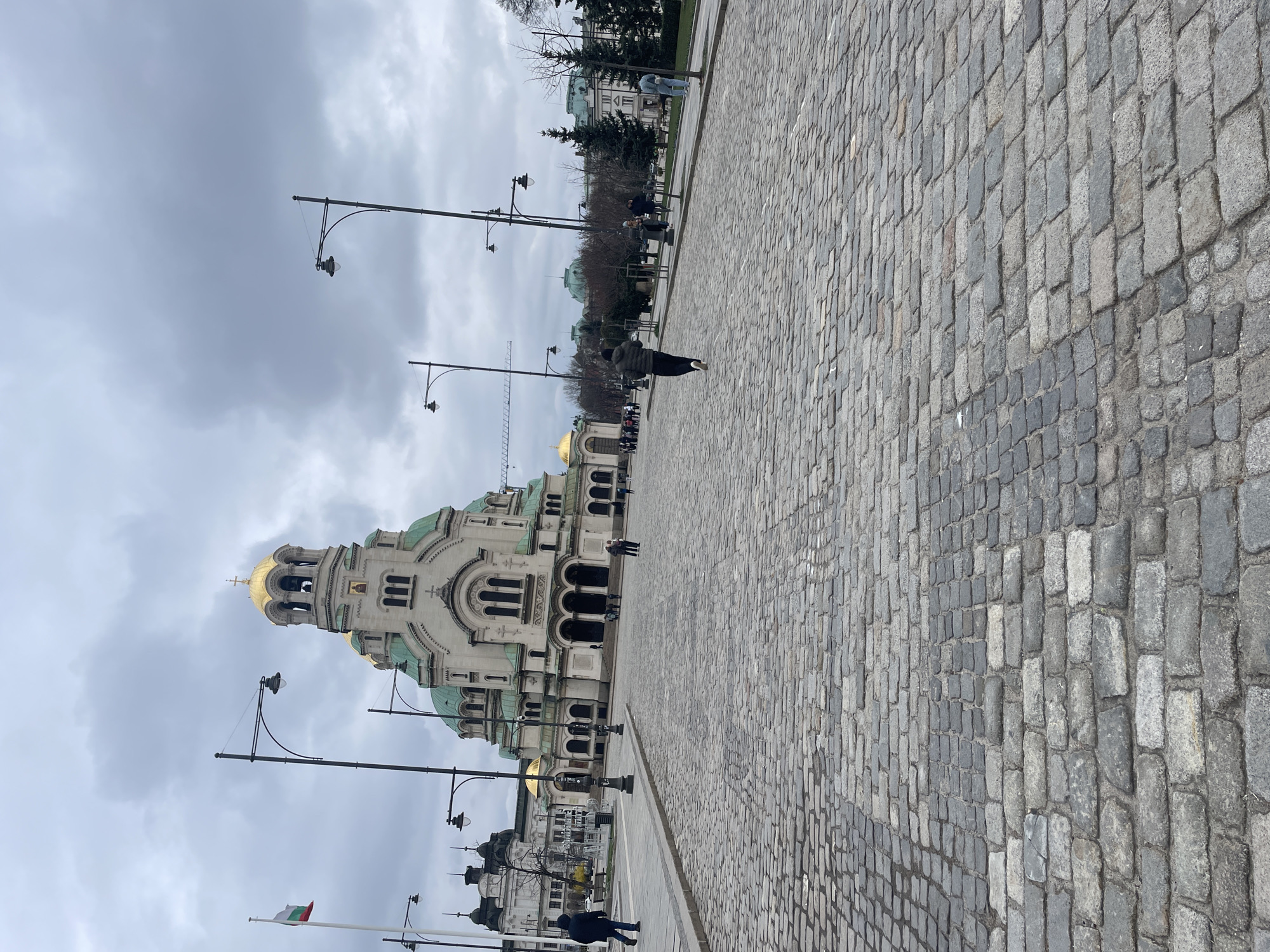The image shows a view of the Alexander Nevsky Cathedral in Sofia, Bulgaria. The cathedral is a large, ornate structure with gold domes and green roofs. The sky is overcast with gray clouds. In the foreground, there is a cobblestone street. People are walking along the street. Street lamps line the street. A Bulgarian flag is seen in the lower-left.