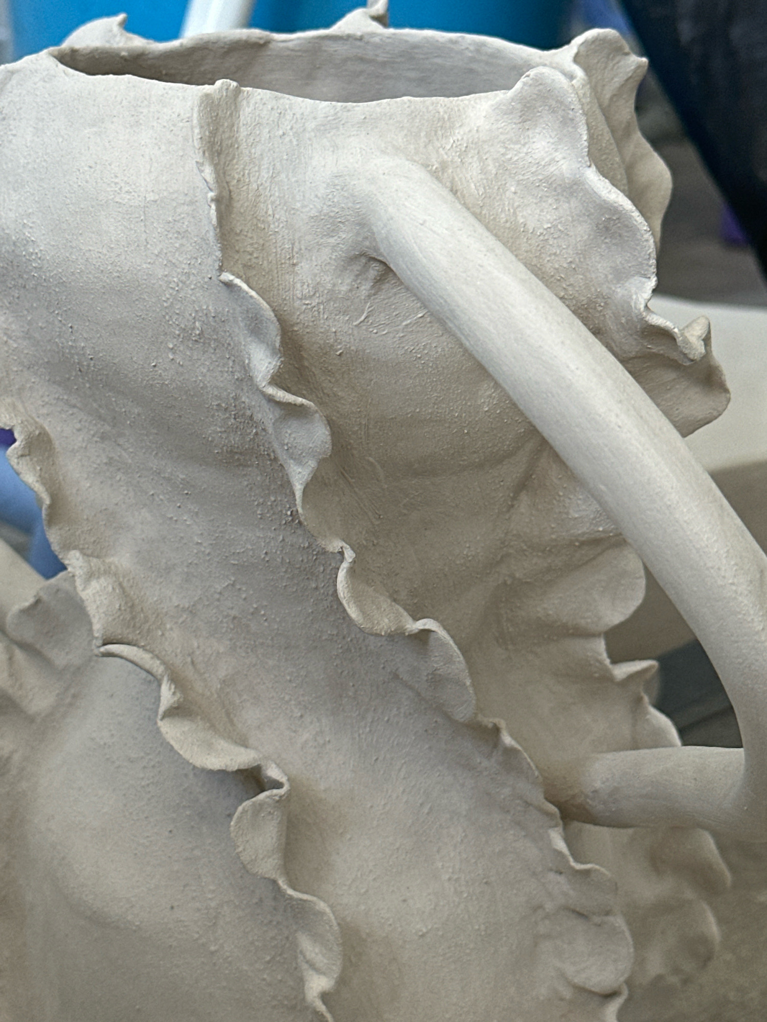 This is a close-up, eye-level, indoor shot of a light-colored, textured ceramic object. The object appears to be a sculpture, possibly a vase or a decorative piece, with ruffled or petal-like edges. A smooth, cylindrical rod-like element is attached to the sculpture, extending diagonally across the frame. The lighting is even, with no harsh shadows. The background is blurred and appears to be a domestic or studio setting with hints of blue and darker colors. There are no people or discernible actions in the image. The location in Nevinnomyssk, Russia cannot be determined from the visual information provided.