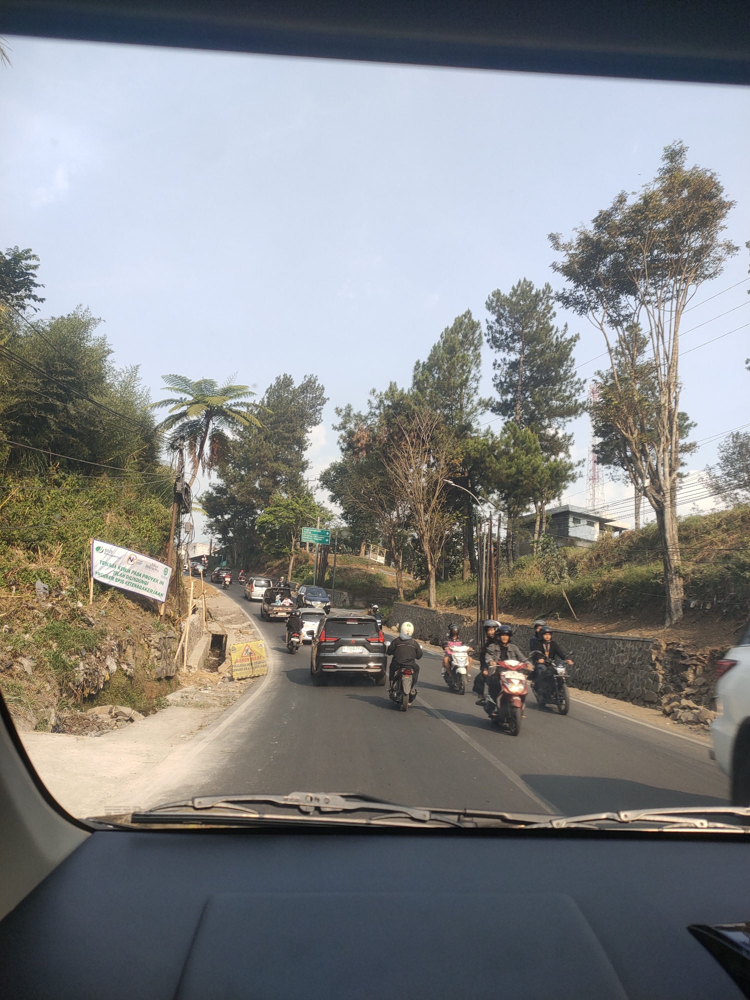 The image depicts a road scene in Cibogo, Indonesia, captured from inside a vehicle. The road is paved and appears to be a two-lane street with traffic, including cars and numerous motorcycles. On the left side of the road, there is a steep embankment covered in vegetation and some litter. A white banner with green text is displayed on this embankment. Further up the embankment, there are trees and a large fern. A utility pole with wires runs alongside the road.

The road itself has several vehicles in motion. Ahead, there are several motorcycles with riders, and a few cars. One car, a black SUV, is in the foreground on the right lane. To the left of the road, there's a construction area with a yellow warning sign.

On the right side of the road, there is a stone retaining wall topped with more vegetation and trees. In the background, there are more trees, some buildings, and a tall red and white tower, possibly a communication tower. The sky is clear and bright. The image appears to have been taken during daylight.