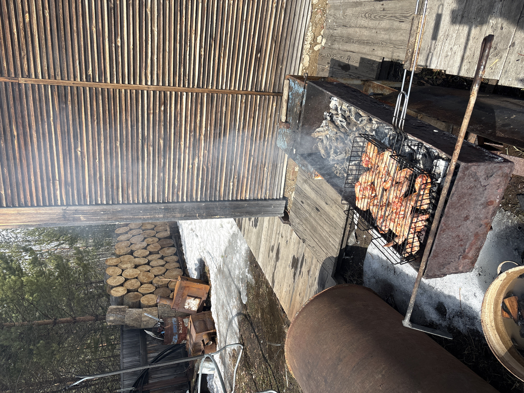 The image depicts a barbecue scene in what appears to be a backyard or outdoor area, possibly located in a rural or suburban setting.
**Objects:**
* A barbecue grill with meat being cooked on a wire rack over hot coals.
* Grilling tools
* Wood logs stacked to the side.
* A metal pipe or cylindrical object.
* Wooden planks making up a deck or platform.
* A shed-like structure with wooden slat walls.
* A bucket containing water and wooden scraps
**Environment:**
* The presence of snow suggests a winter or early spring season.
* Trees visible in the background indicate a location near a wooded area.
Given the request to include the location in Zheleznodorozhnyi, Russia, it is possible that this scene is occurring in the area surrounding this city, possibly in a dacha or private property outside the urban center.