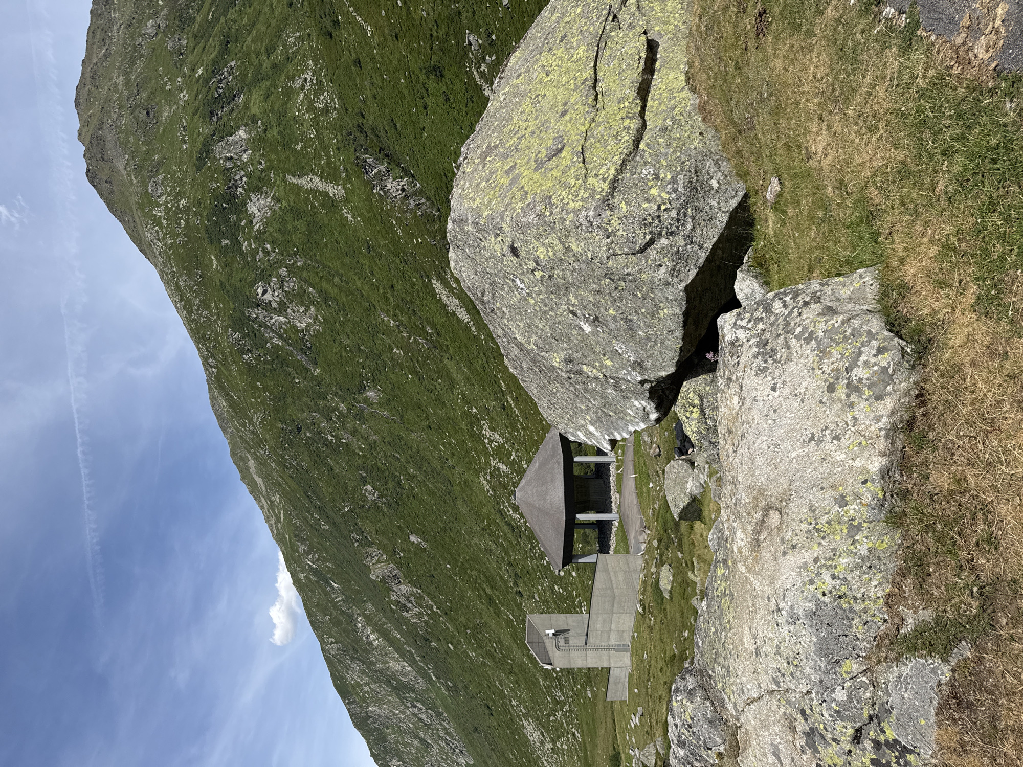 In Hospental, Switzerland, a mountain landscape is depicted under a partly cloudy sky. In the foreground, large, lichen-covered boulders rest on a grassy slope. Behind them, a modern concrete structure stands to the left, featuring a vertical tower with antennas and a lower, angled section. To the right of the concrete building, an open-sided pavilion with a thatched roof is situated on a paved area. The background is dominated by a steep, green mountain slope dotted with rocks and vegetation. A thin contrail is visible in the sky.
