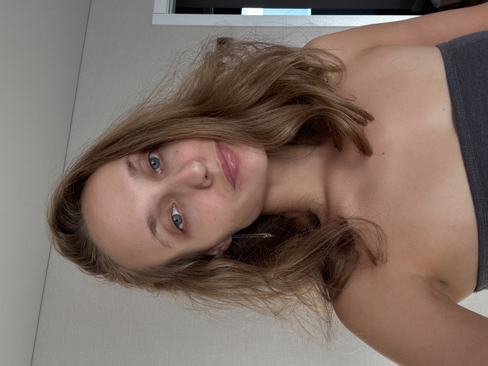The image shows a close-up, head-and-shoulders portrait of a young woman with light brown, wavy hair, fair skin, and blue eyes. She is wearing a dark gray strapless top and a delicate silver chain earring that dangles from her left ear. Her expression is neutral, with slightly parted lips. The background appears to be an indoor setting, with a light-colored, textured wall and a doorway visible on the right. The lighting suggests it is daytime. There are no observable elements that would indicate a specific location within the city of Подшивалово, Russia.