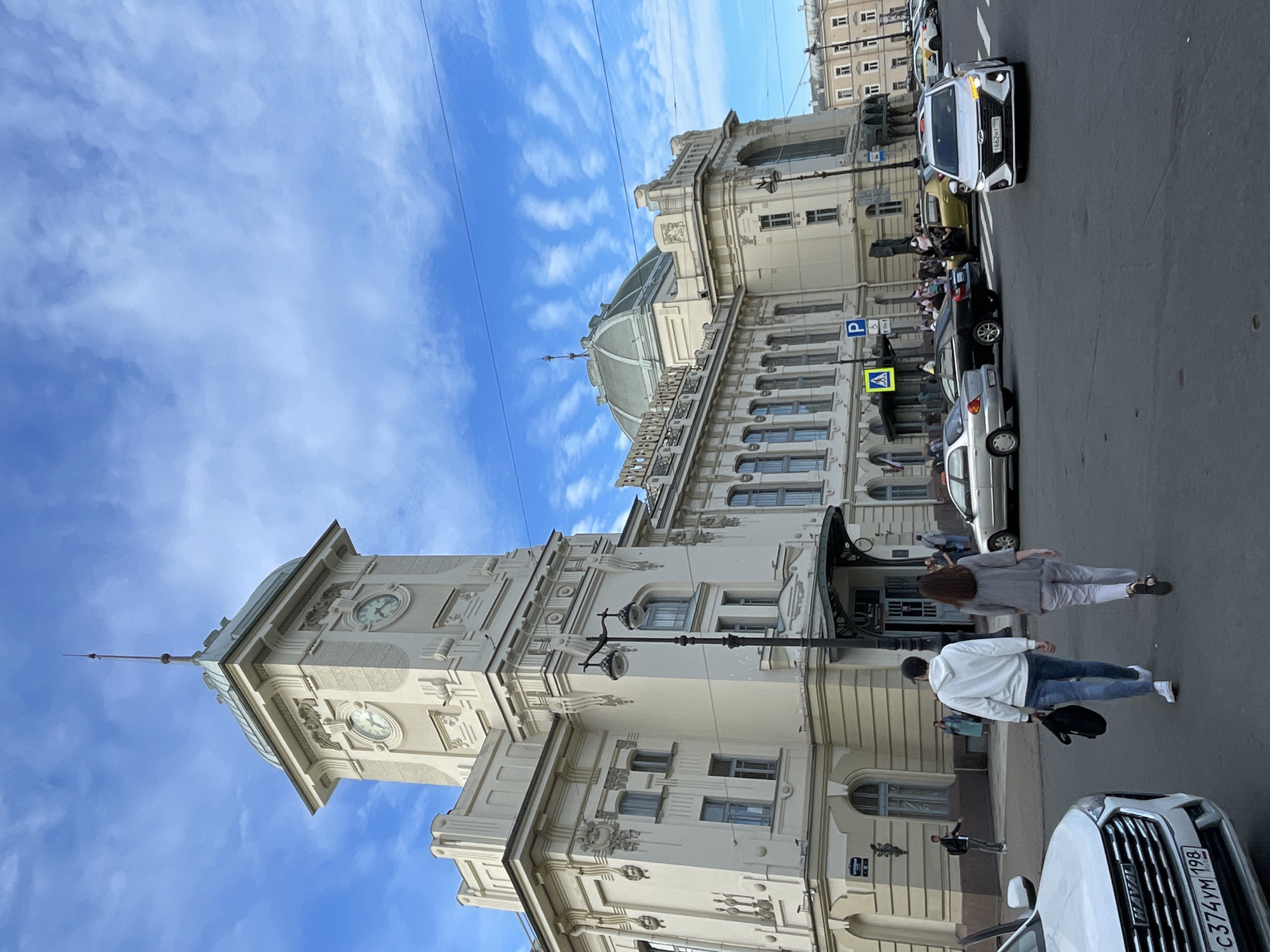 The image captures a street-level view of the Vitebsky Railway Station in Saint Petersburg, Russia, on a day with a partly cloudy sky. The grand, cream-colored station building dominates the background, featuring a prominent clock tower with a green domed roof and ornate architectural details. The street in the foreground is paved, with several cars present. A white Haval SUV is parked on the left side of the frame, its license plate visible. Two individuals, a man in a white hoodie and jeans and a woman in a grey top and trousers, are walking away from the camera towards the station entrance. Other pedestrians are visible near the entrance and on the sidewalks. Several cars are in motion or parked on the street, including a silver sedan, a black sedan, and a white sedan with its headlights illuminated. Traffic signs, including a parking sign and a pedestrian crossing sign, are also visible. The overall scene depicts an active urban environment with a mix of historical architecture and contemporary life.