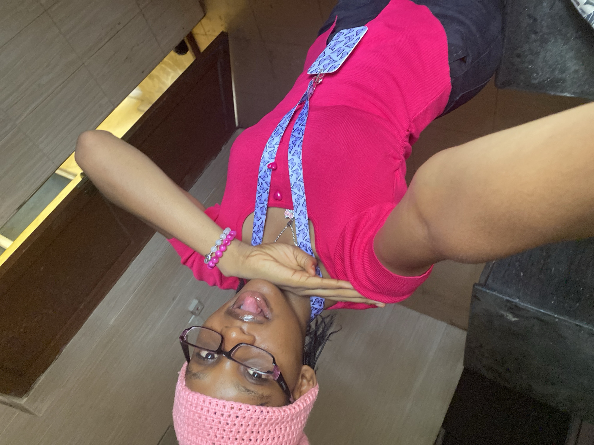 A person wearing glasses and a pink crocheted headband is taking a selfie indoors, possibly in a bathroom. They are wearing a pink shirt with a butterfly lanyard and a bracelet. They are sticking out their tongue. The location is the city of Unknown, Unknown.