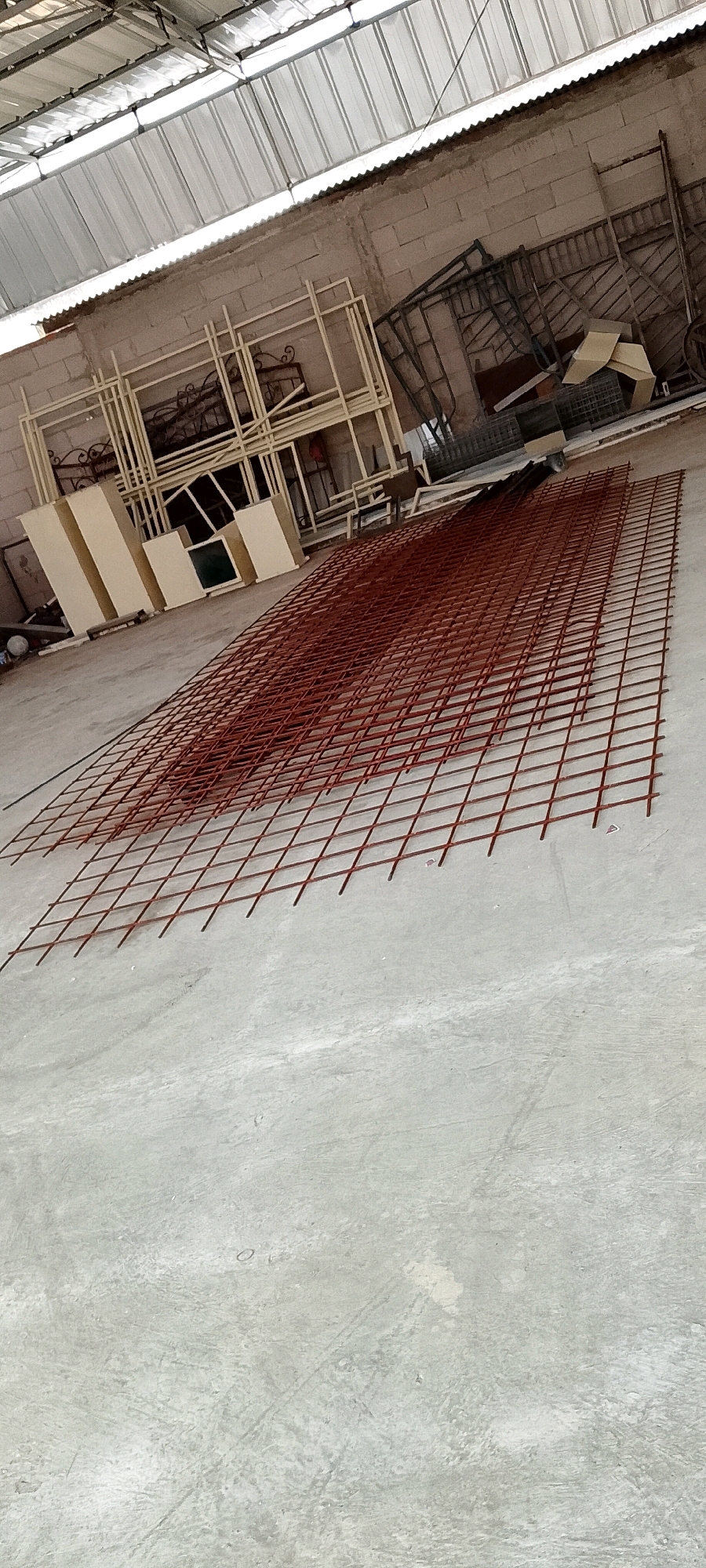 An indoor scene depicts a workshop or storage area, likely in Sriamur, Indonesia. A large, rectangular grid of reddish-brown metal mesh lies on a concrete floor, extending from the foreground into the mid-ground. Behind the mesh, several light-colored cabinets or storage units are arranged against a pale brick wall. Further back, various metal frames and structural elements are visible, stacked and leaning. The ceiling is constructed of corrugated metal panels, allowing natural light to filter into the space. The overall impression is one of a functional, utilitarian environment.