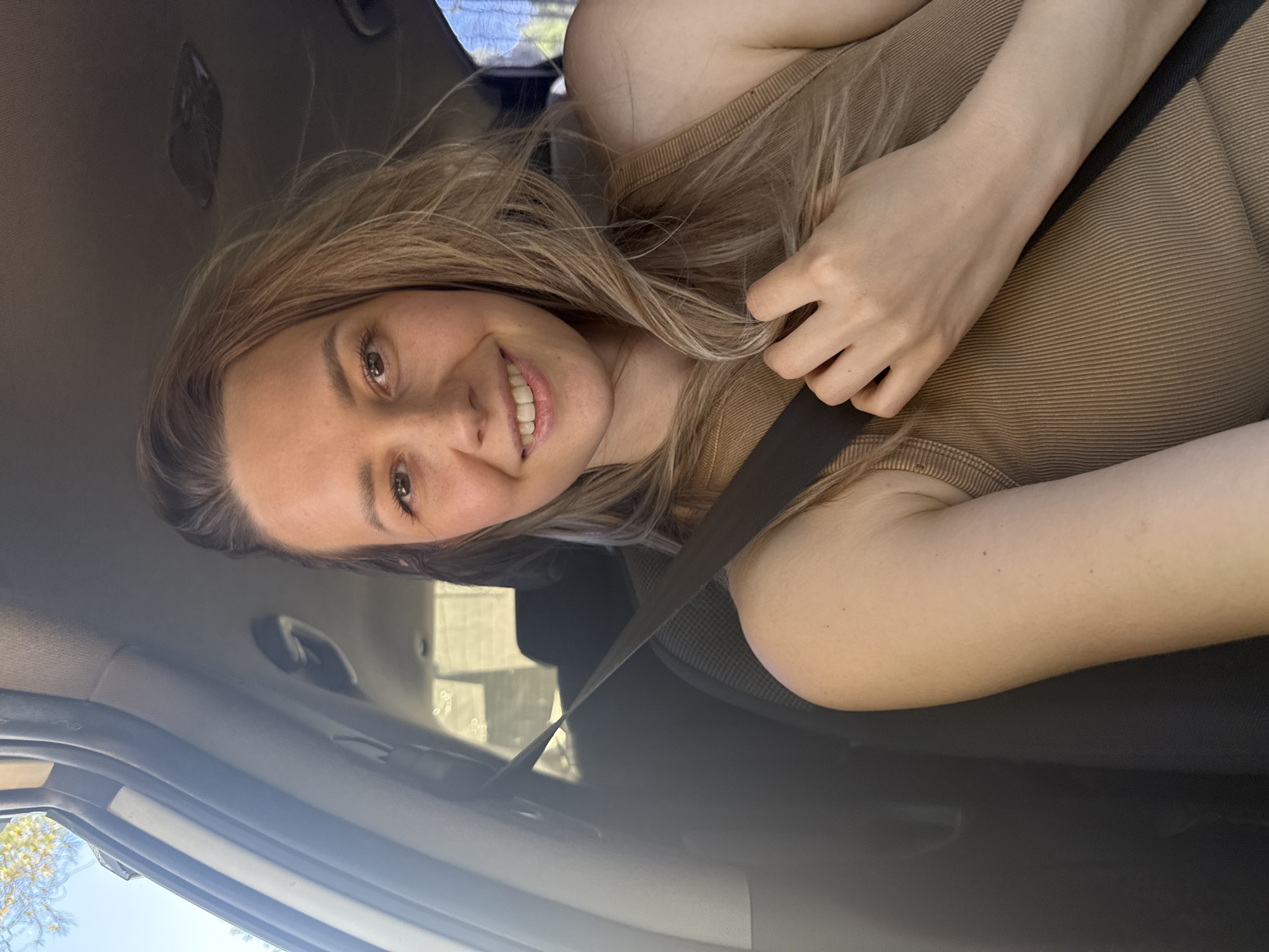 A woman with long blonde hair, wearing a brown top and a seatbelt, is sitting in a car. The car is likely parked, based on the presence of the seatbelt and the lack of motion blur. The location is unknown and there is no indication of it being in Chaikovsky, Russia.