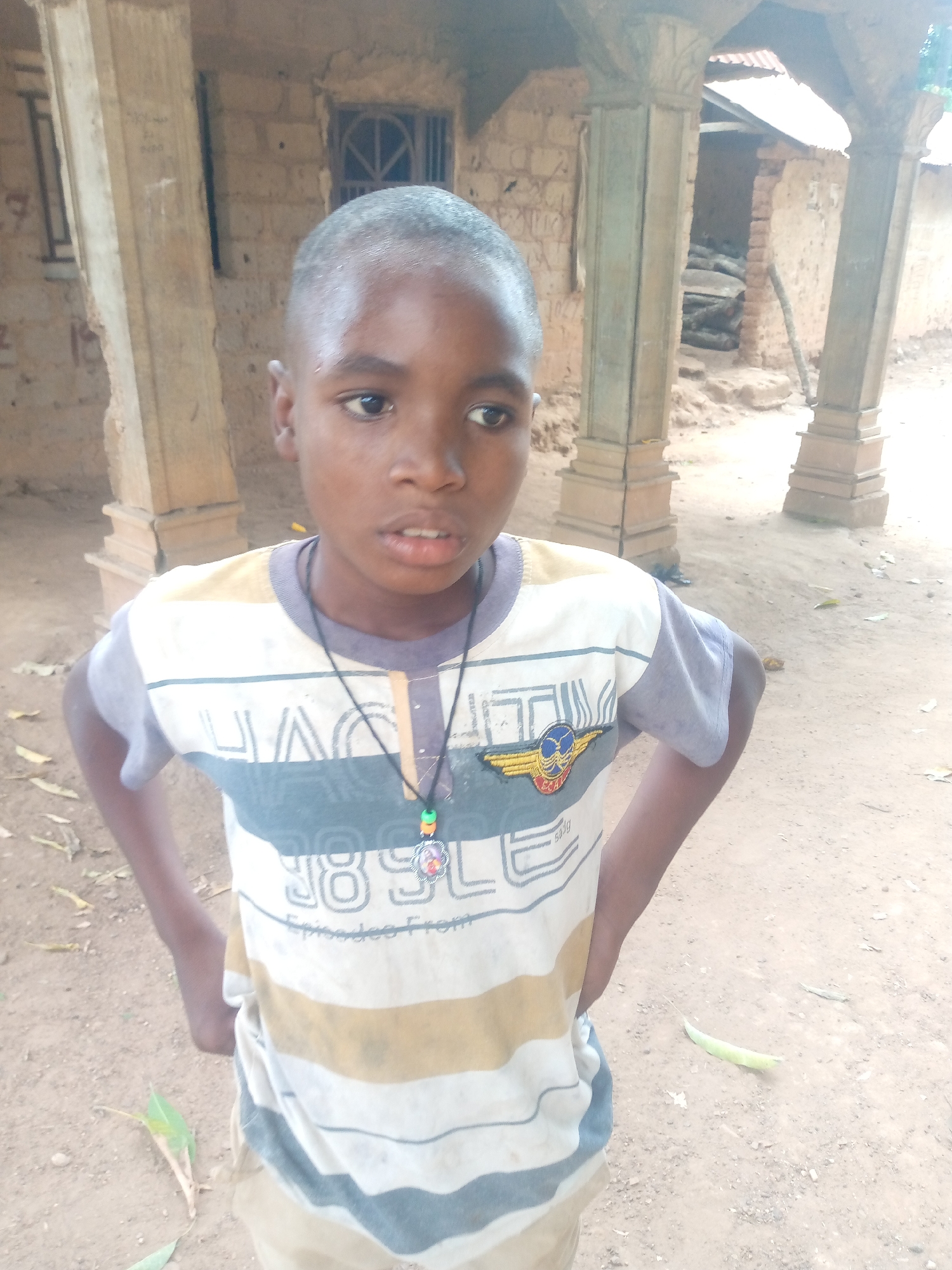 A young Black boy stands outdoors, his hands on his hips. He wears a short-sleeved striped shirt with a pendant necklace. He has a closely shaved head. Behind him is a structure supported by pillars. The ground is dirt. The location is the city of Akwanga, Nigeria.