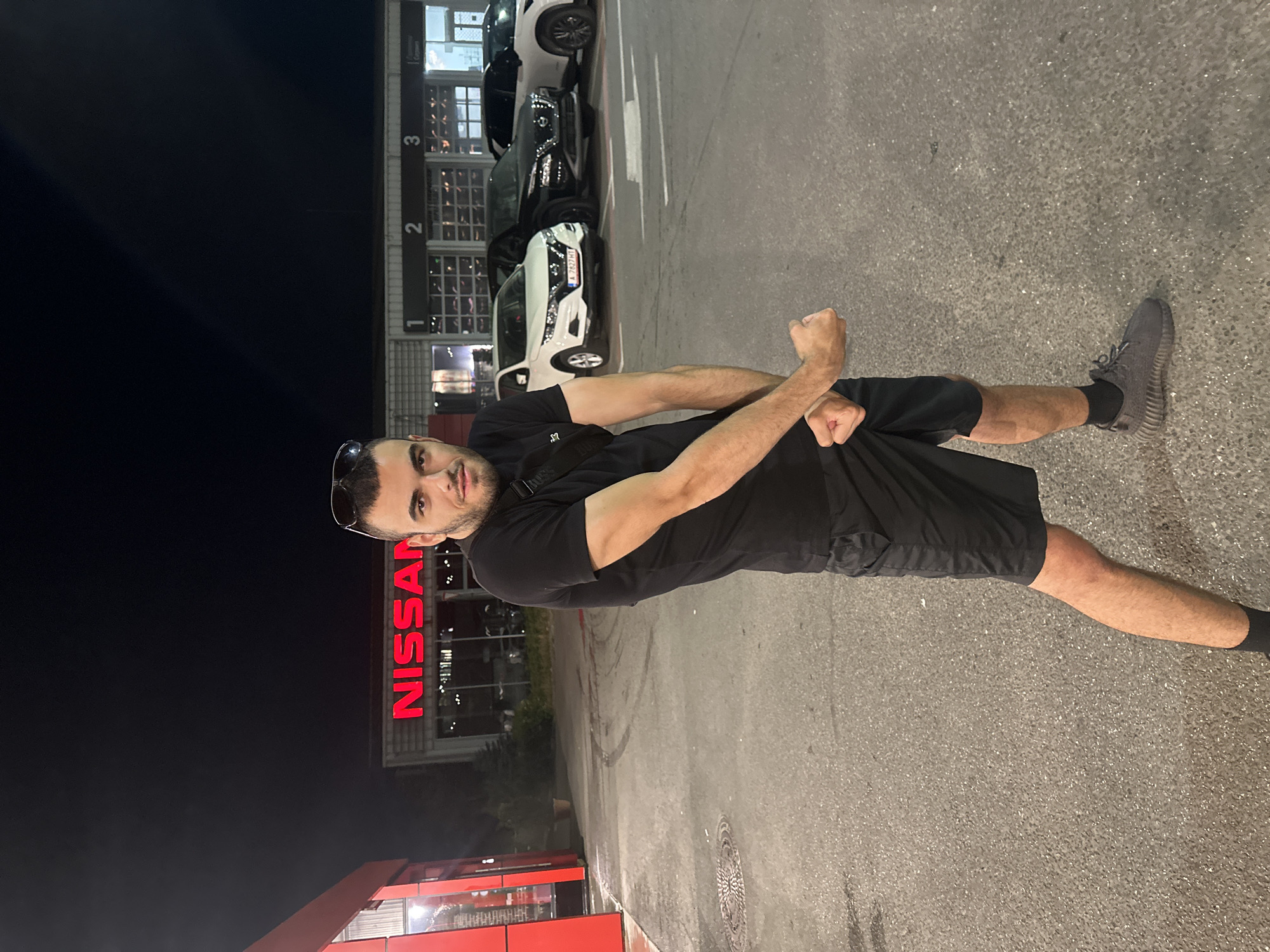 A full-body, eye-level shot shows a man posing outdoors at night in front of a Nissan dealership located in Burgas, Bulgaria. The man, who appears to be in his early twenties, has short dark hair and a beard. He is wearing a black t-shirt, black shorts, dark grey sneakers, and a black fanny pack across his chest. He is flexing his biceps, and has sunglasses perched on his head. Behind him, the Nissan dealership has large, illuminated red "NISSAN" lettering and several vehicles parked outside. The ground is asphalt, and there are tire marks visible. The scene is lit by artificial lighting, creating a nighttime ambiance.