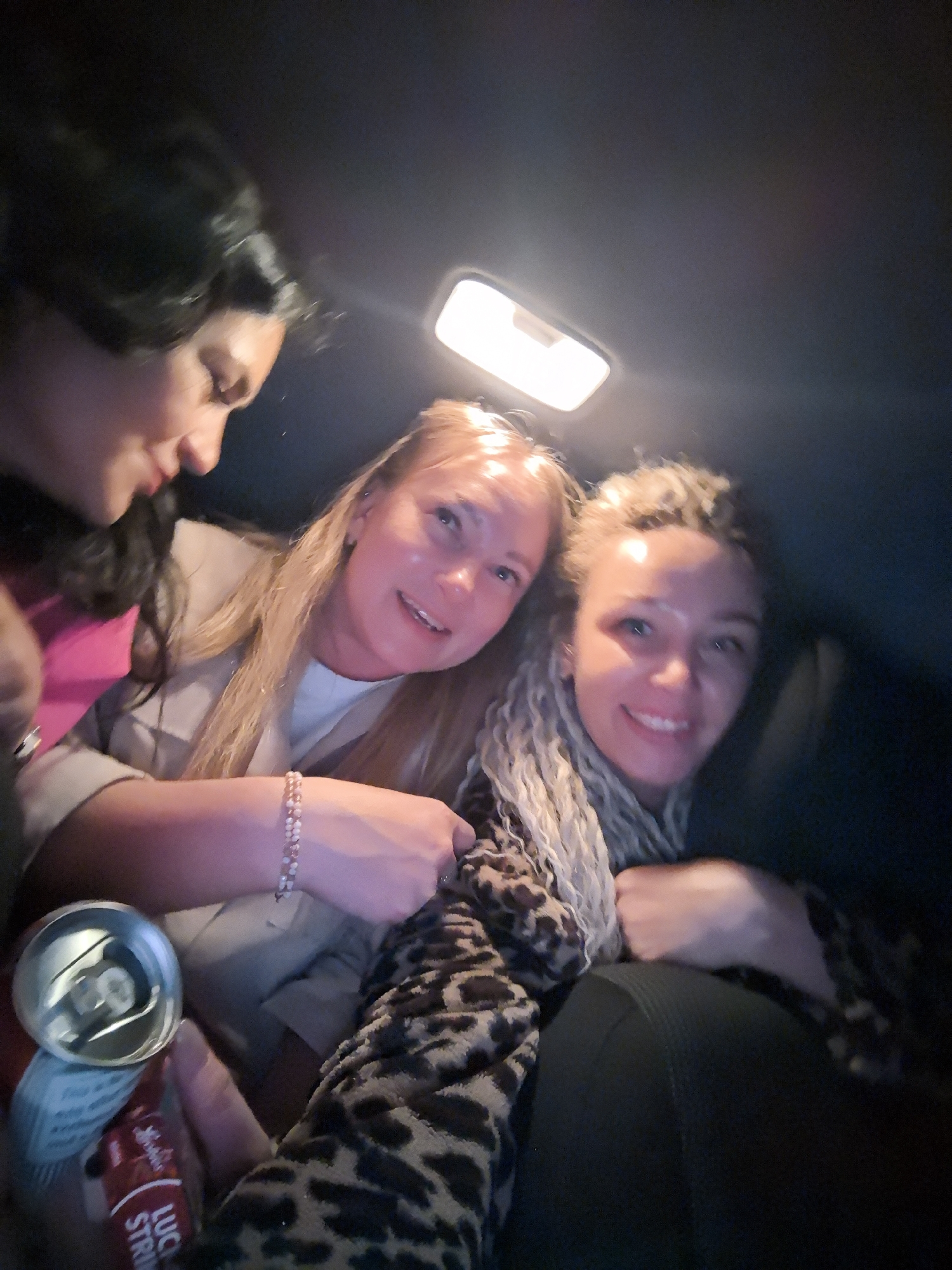 Three women are in a dimly lit vehicle, likely a car, based on the interior light fixture visible overhead. The woman on the left has dark hair and wears a pink top; she is looking down and to the right. The woman in the center has blonde hair and wears a light-colored blazer over a white shirt; she is smiling and looking towards the camera. The woman on the right has blonde dreadlocks and is wearing a leopard-print jacket over a dark shirt; she is also smiling and looking at the camera. One person in the group is holding an open can of "Lucky Strike" brand beverage. The location is unclear, but the user suggests that it is the city of Noviy, Russia. There is no additional verifiable information.