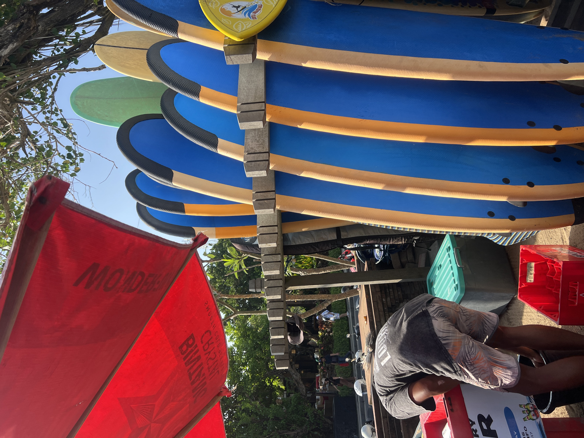 In Kuta, Indonesia, a person wearing a gray t-shirt and patterned shorts is bent over, rummaging through a red crate. The scene is set outdoors, with a red awning overhead, possibly belonging to a beachside establishment. A row of surfboards, predominantly blue with yellow accents, stands upright, secured by a wooden rack. Behind the surfboards, several other surfboards of varying colors, including green and yellow, are visible, along with trees and a clear blue sky. In the background, people are walking along a street, and a motorcycle is parked. Various containers, including a green plastic bin and a red plastic crate, are scattered around the immediate area. The ground appears to be sandy or dusty.