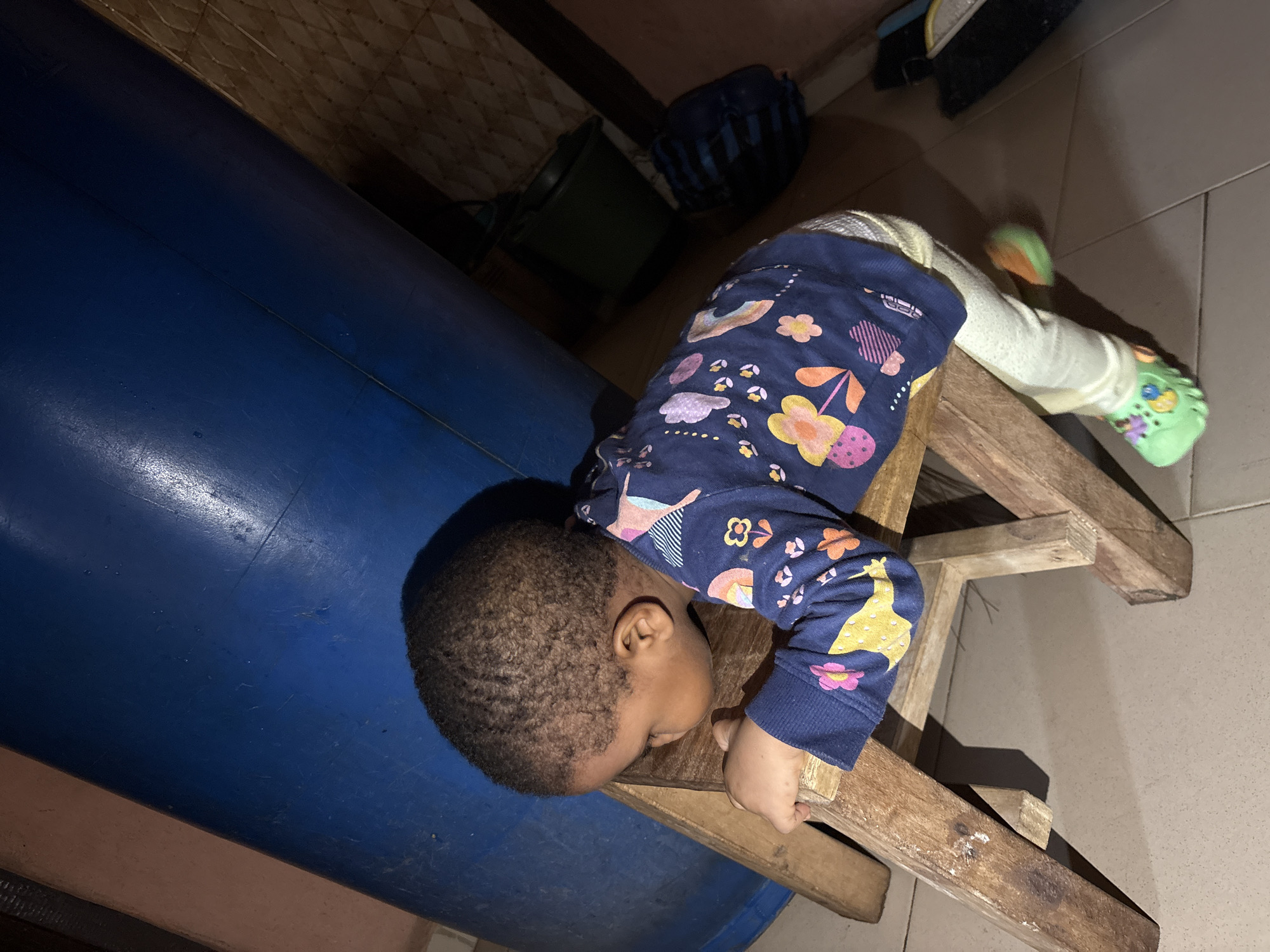 A young child with dark, short hair, wearing a dark blue long-sleeved shirt with a colorful floral pattern and light-colored leggings, is leaning over a wooden stool. The child's left hand rests on the stool's seat, and their head is turned away from the camera. The child is wearing green slip-on shoes with colorful embellishments. The stool is made of unfinished wood and has a simple, rustic design with four legs. In the background, a large, blue cylindrical object, possibly a water tank, dominates the upper portion of the image. To the right of the blue object, a dark green plastic bucket is visible, and further to the right, a dark blue duffel bag rests on the floor. The floor appears to be tiled with a light brown diamond pattern. The wall behind the bucket and duffel bag has a textured surface with a diamond pattern. The scene is lit with artificial light, casting shadows on the floor and the stool. This image was taken in Onitsha, Nigeria.