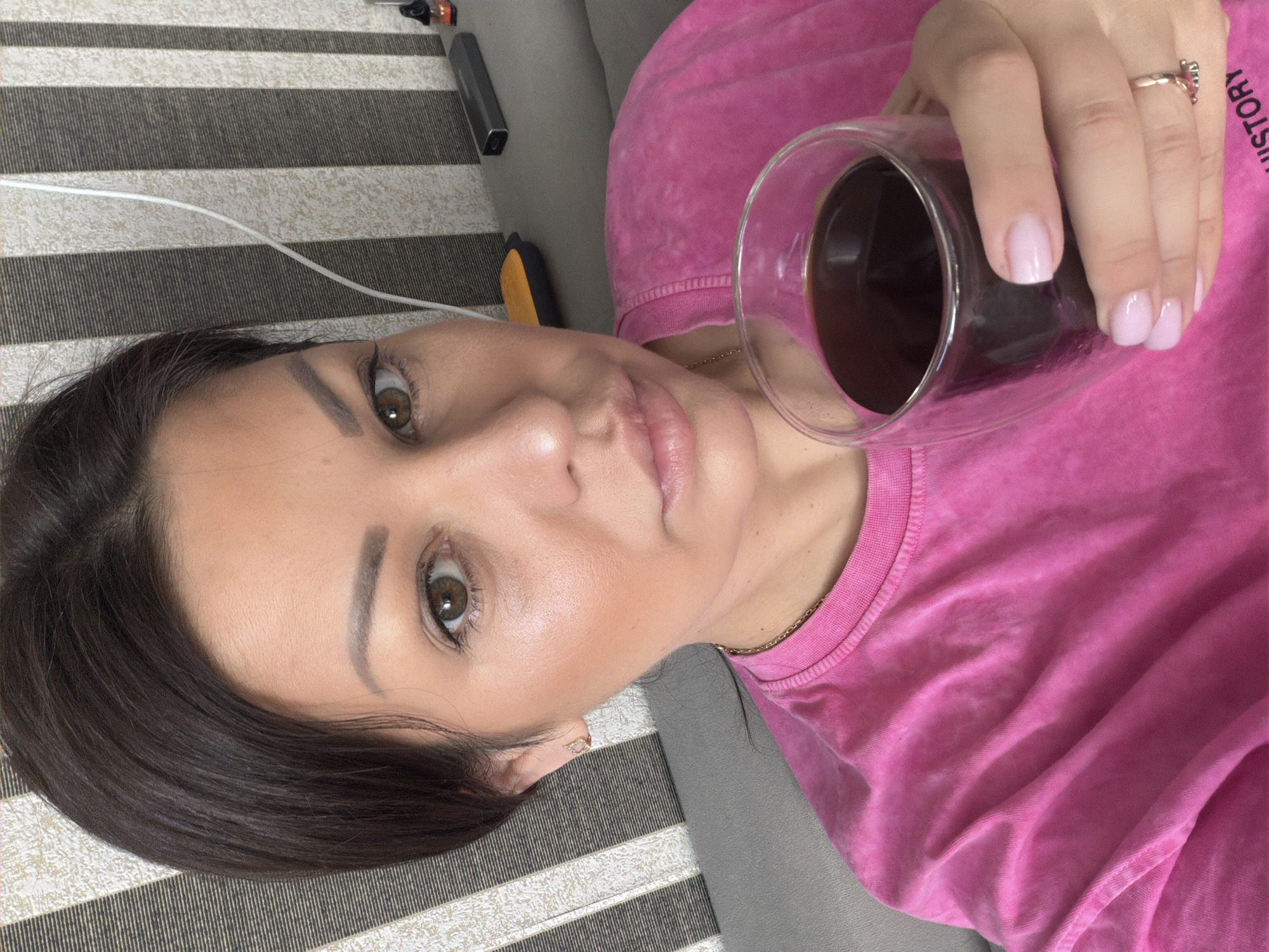 A medium shot captured from a slightly high angle shows a woman holding a glass of dark liquid, presumably coffee, in her right hand. She is seated on a gray sofa, with a striped wallpaper pattern visible in the background. Her hair is dark brown and pulled back, revealing her face which is partially illuminated. She is wearing a bright pink t-shirt and a thin gold necklace. The woman's gaze is directed towards the camera. A ring adorns her right ring finger, and her fingernails are painted a pale pink. The overall setting appears to be an indoor domestic environment, possibly a living room, in Sochi, Russia.